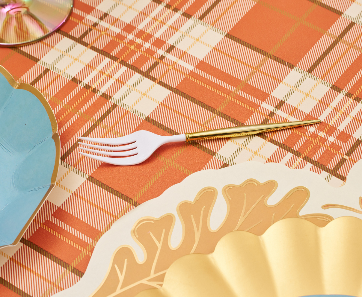 A white and gold fork rests on an orange plaid tablecloth near blue and gold scalloped plates from the Turkey & Gourds: Thanksgiving Table for 16 collection by Sophistiplate LLC, creating an inviting Thanksgiving tablescape.