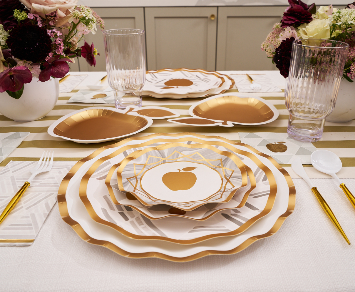 Create a stunning Rosh Hashanah tablescape with the Sophistiplate Golden Apple Table Setting, featuring gold-rimmed disposable plates adorned with a gold apple motif, complemented by gold utensils, clear glasses, and floral centerpieces on a striped runner—everything you need for an elegant celebration.