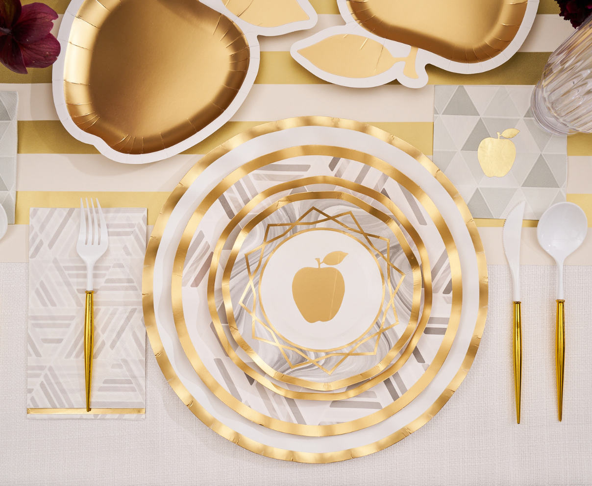 Introducing the Sophistiplate Golden Apple Table Setting, ideal for Rosh Hashanah celebrations. This elegant set features gold and white disposable plates decorated with an apple motif, each place set complete with sparkling gold utensils, a neatly folded napkin, and sophisticated gold-accented apple-shaped plates arranged on a chic striped placemat.