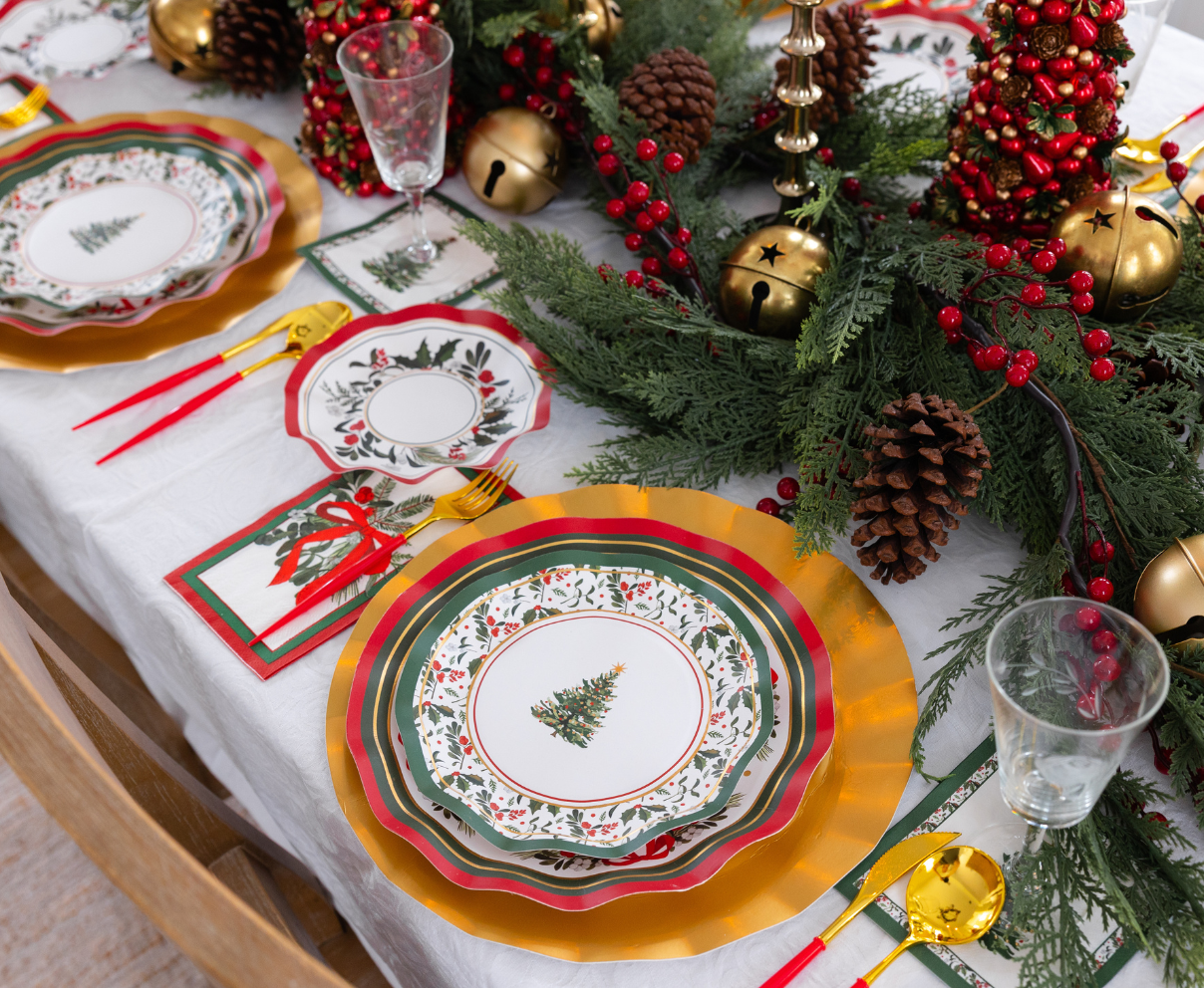 A Home for the Holidays: Christmas Table for 16 setting featuring heavy duty paper plates with Christmas tree designs layered on gold chargers, with red and gold cutlery, napkins, and a centerpiece of pine branches, pinecones, and red berries.