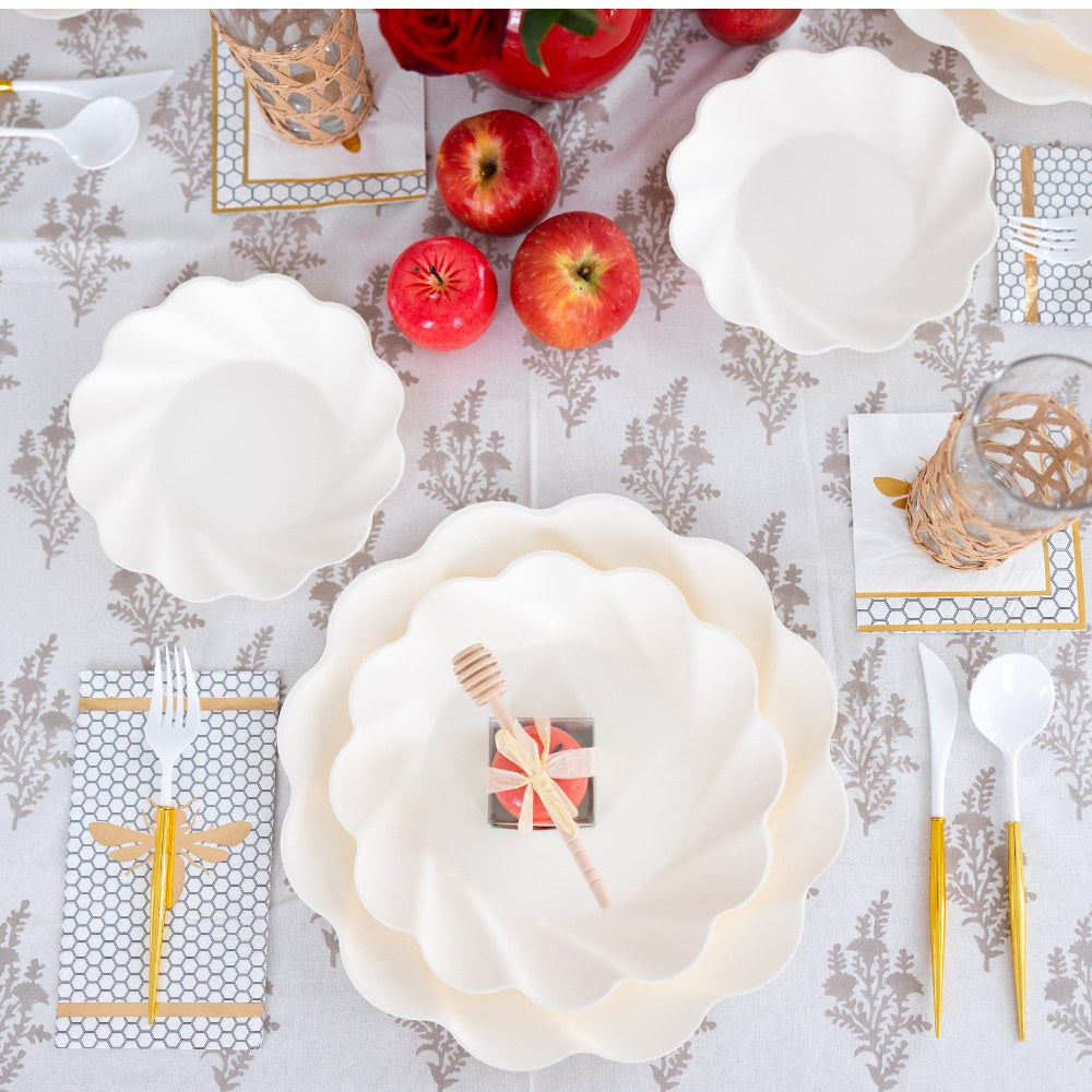 The table is set with Sophistiplate LLC’s “Rosh Tablescape for 16 Guests: Eco Cream Honeybee,” featuring elegant white scalloped disposable plates and bowls, apples, a honey dipper placed on a small gift box, glasses with wicker sleeves, and gold utensils arranged on a leaf-patterned tablecloth.