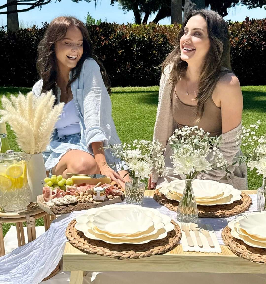 Two women smile and relax on a picnic blanket in the park, enjoying snacks and drinks served with Sophistiplate’s Eco-Friendly Table Setting- Cream, surrounded by flowers on a low wooden table during a sunny outdoor day.