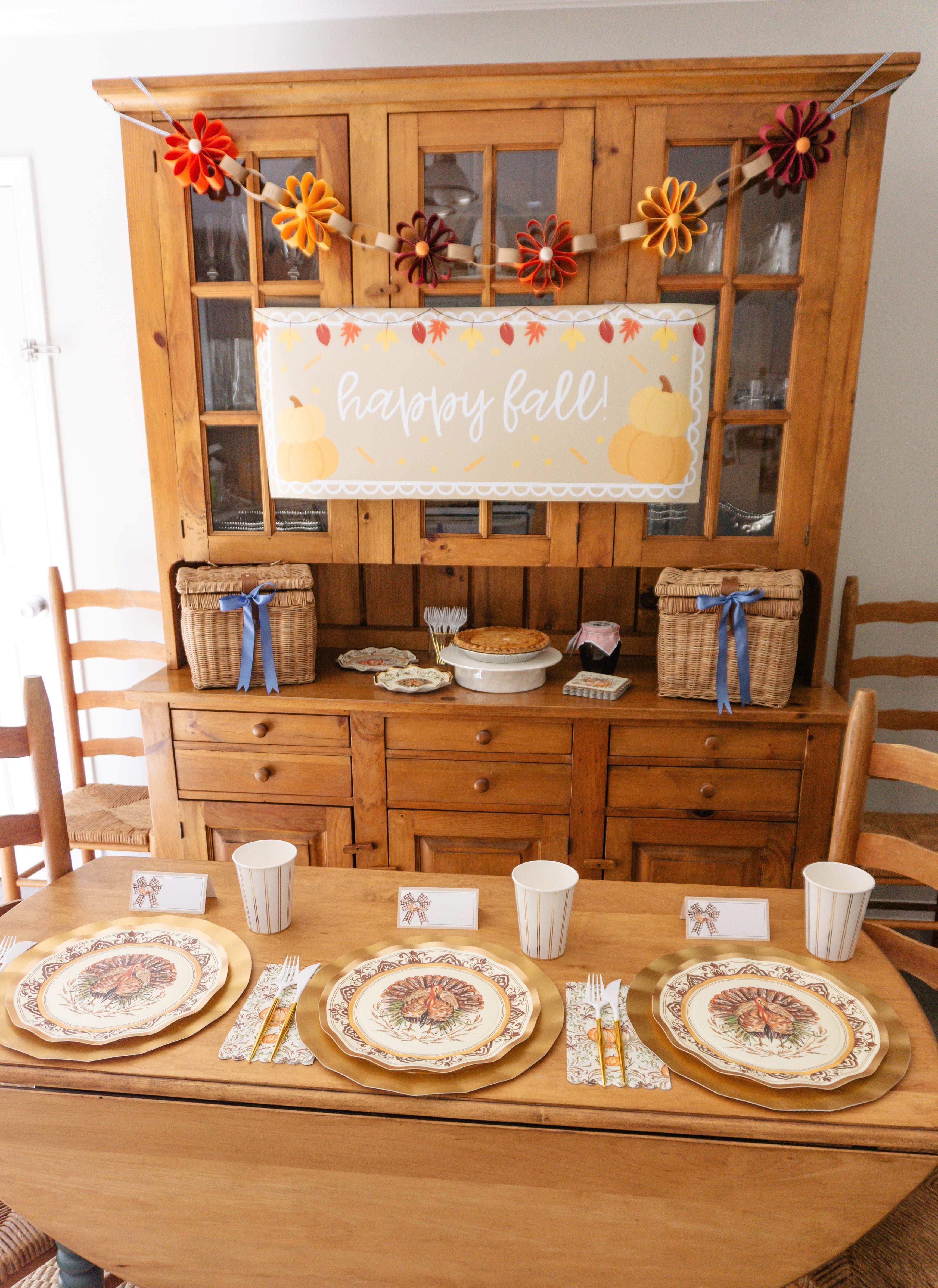 A Thanksgiving table setting featuring the Turkey & Gourds: Thanksgiving Table for 16 set with heavy-duty paper plates, gold chargers, and cups, with a happy fall banner hung on the hutch behind the table.
