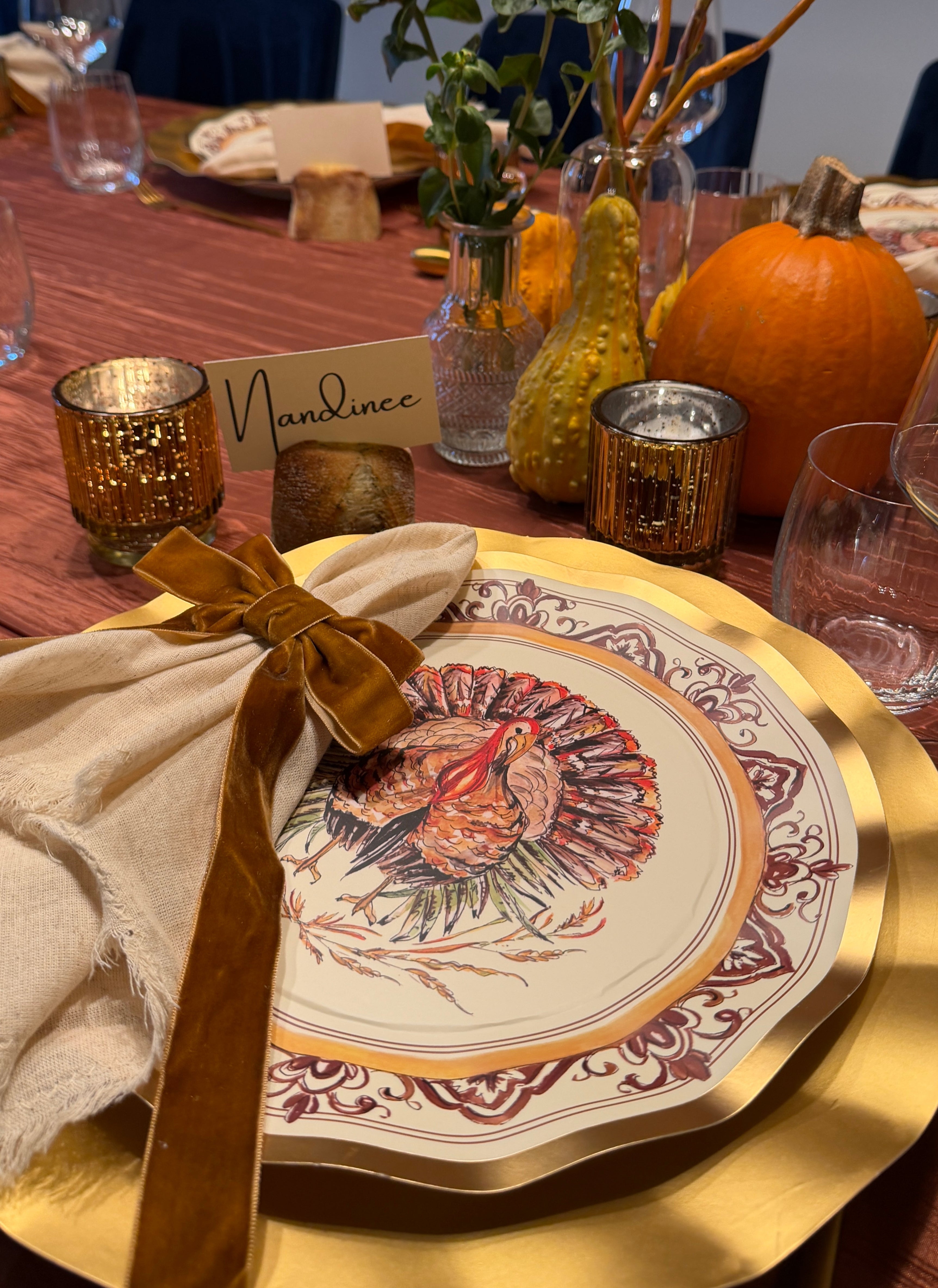 A Turkey & Gourds: Thanksgiving Table for 16 setting featuring a heavy duty paper plate with a turkey illustration, a beige napkin tied with a brown ribbon, and a place card labeled Nandinee, surrounded by gourds and candles on a red tablecloth.