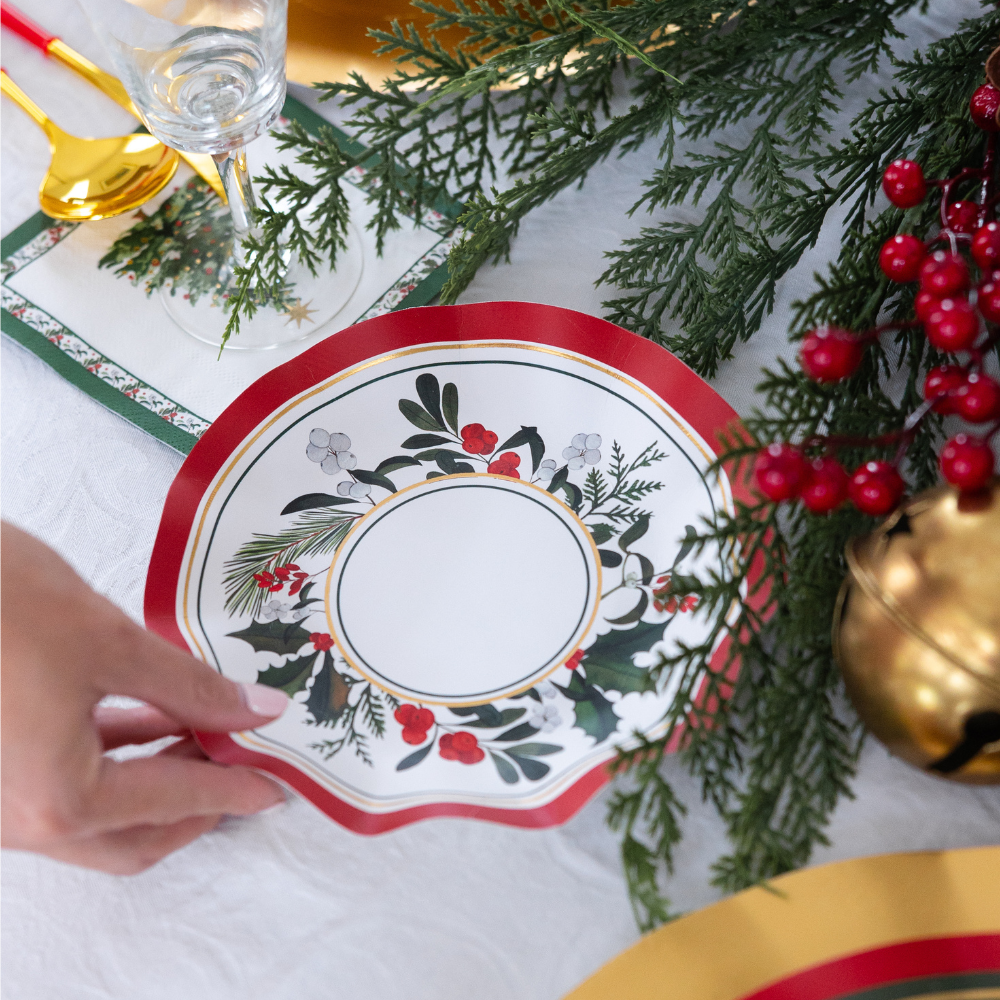 A hand places a Sophistiplate LLC Home for the Holidays: Christmas Table for 16 white scalloped-edge paper plate with a red rim and holly illustrations onto a festive table decorated with evergreen sprigs, red berries, and a gold bell.