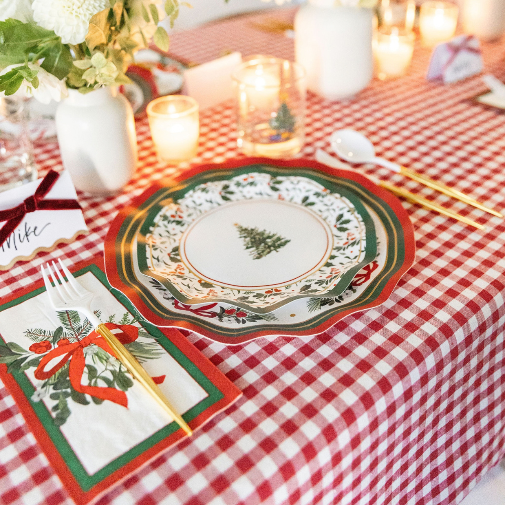 A Home for the Holidays: Christmas Table for 16 place setting on a red and white checkered tablecloth featuring heavy duty paper plates with a pine tree design, a festive napkin, a gold-toned fork and spoon, and a place card with a velvet ribbon.