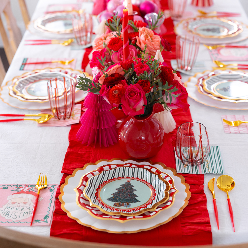 A Tis the Season: Christmas Table for 16 setting with tiered designer paper plates featuring tree illustrations, red napkins with a Santa graphic, gold cutlery with red handles, striped glasses, and a centerpiece of red roses and carnations.