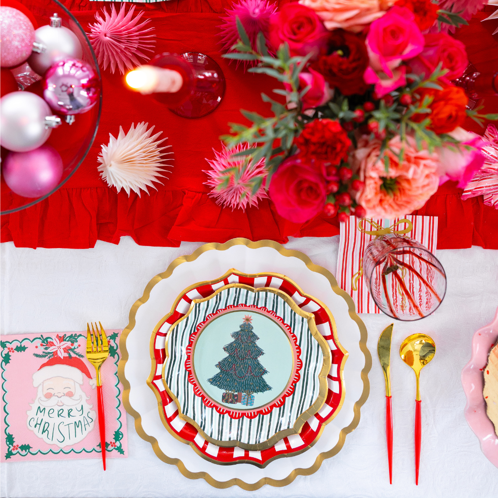 A Tis the Season: Christmas Table for 16 setting featuring designer paper plates with a tree design and a tiered red and white scalloped border, gold-handled cutlery, a festive Santa napkin, and a floral centerpiece on a red runner.