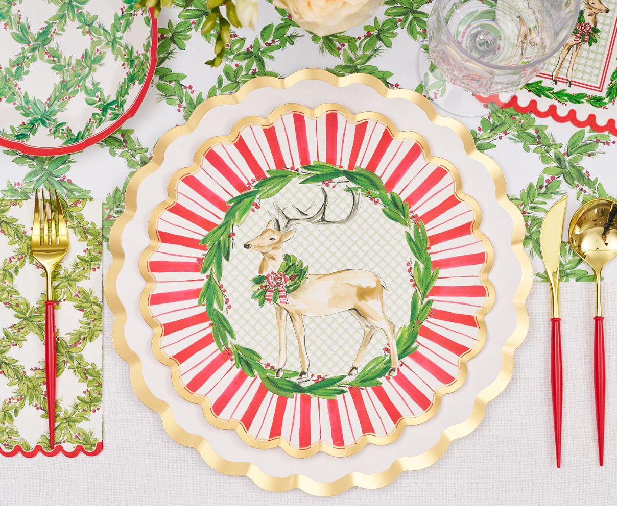 The Sophistiplate Holly Berry Table Setting features gold utensils, reindeer and wreath designer plates, holly napkins, a clear glass, and a white holiday-themed tablecloth for a festive Christmas tablescape.