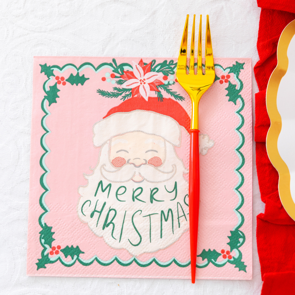 A pink paper napkin with a Santa Claus illustration and Merry Christmas text from the Tis the Season: Christmas Table for 16 set, featuring a gold and red fork next to white and gold designer paper plates.