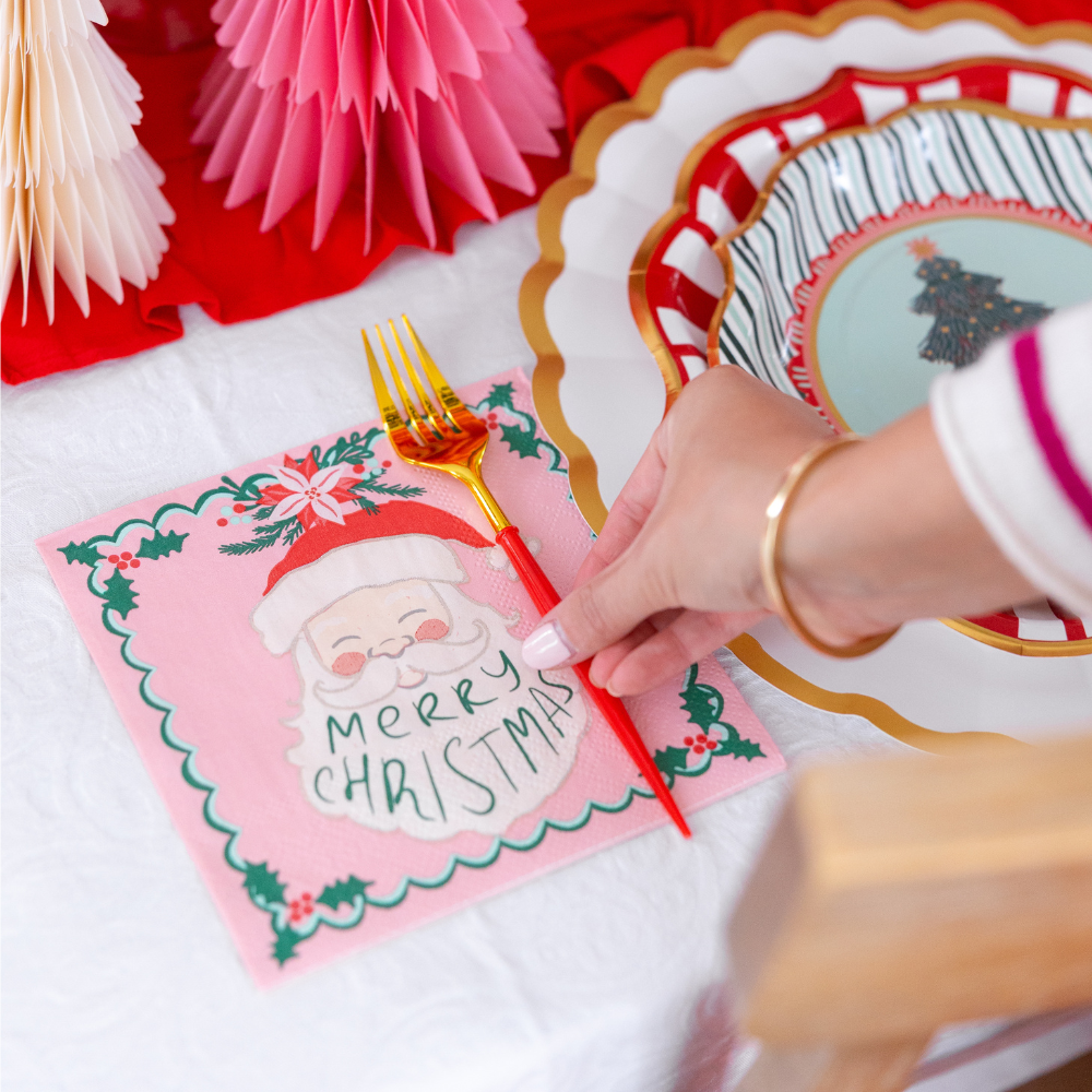 A hand places a red fork onto a Santa Claus napkin next to layered plates from the Tis the Season: Christmas Table for 16 set by Sophistiplate LLC.