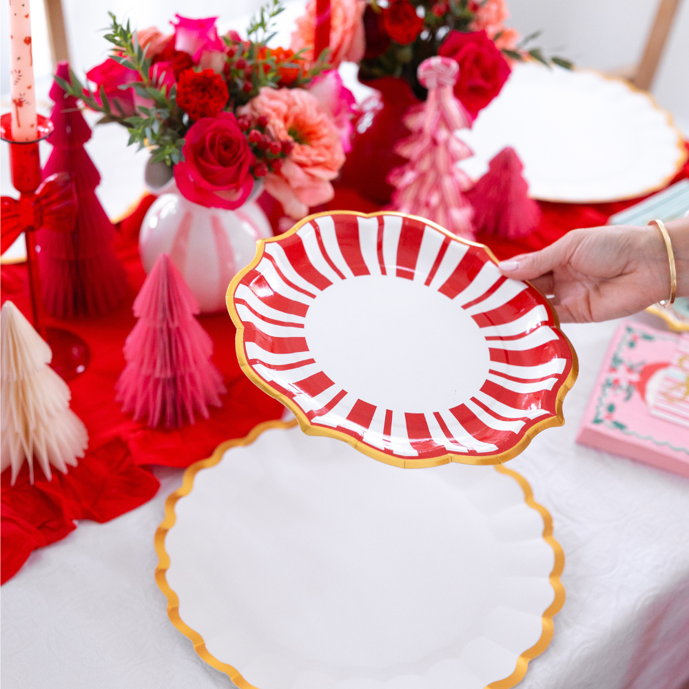 A hand holds a scalloped, red-and-white striped Tis the Season: Christmas Table for 16 designer paper plate over a matching white plate with a gold rim on a table set for Christmas.