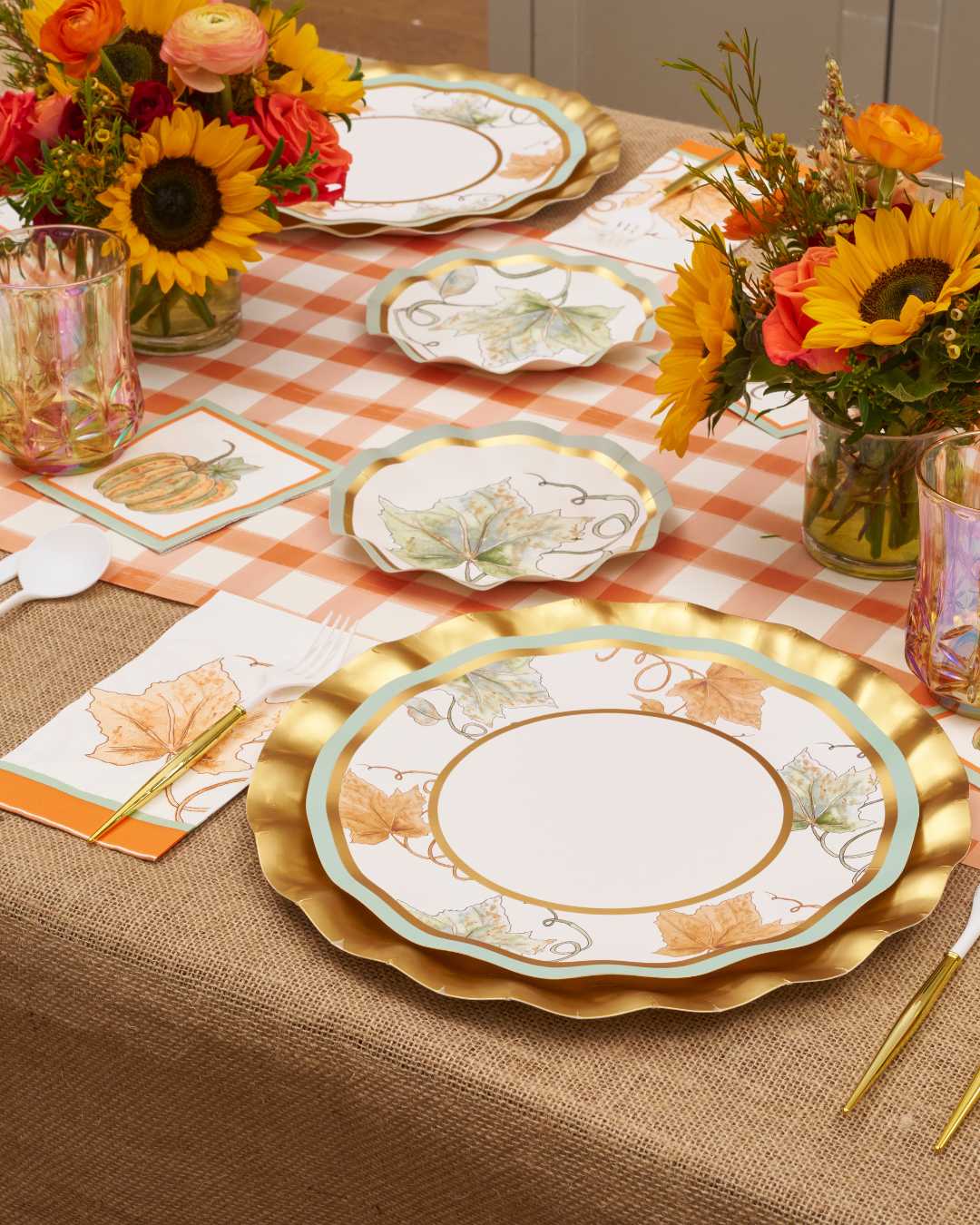 The Bright & Thankful Table Setting by Sophistiplate features gold-rimmed pumpkin plates, floral and leaf motifs, colorful glasses, napkins on plaid burlap, and sunflower centerpieces for a festive autumn table.