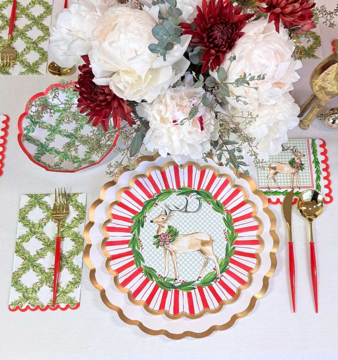 Create a festive holiday tablescape with the Sophistiplate Holly Berry Table Setting, featuring scalloped reindeer plates, gold-and-red utensils, a white and red floral centerpiece, and green-patterned decorative napkins.