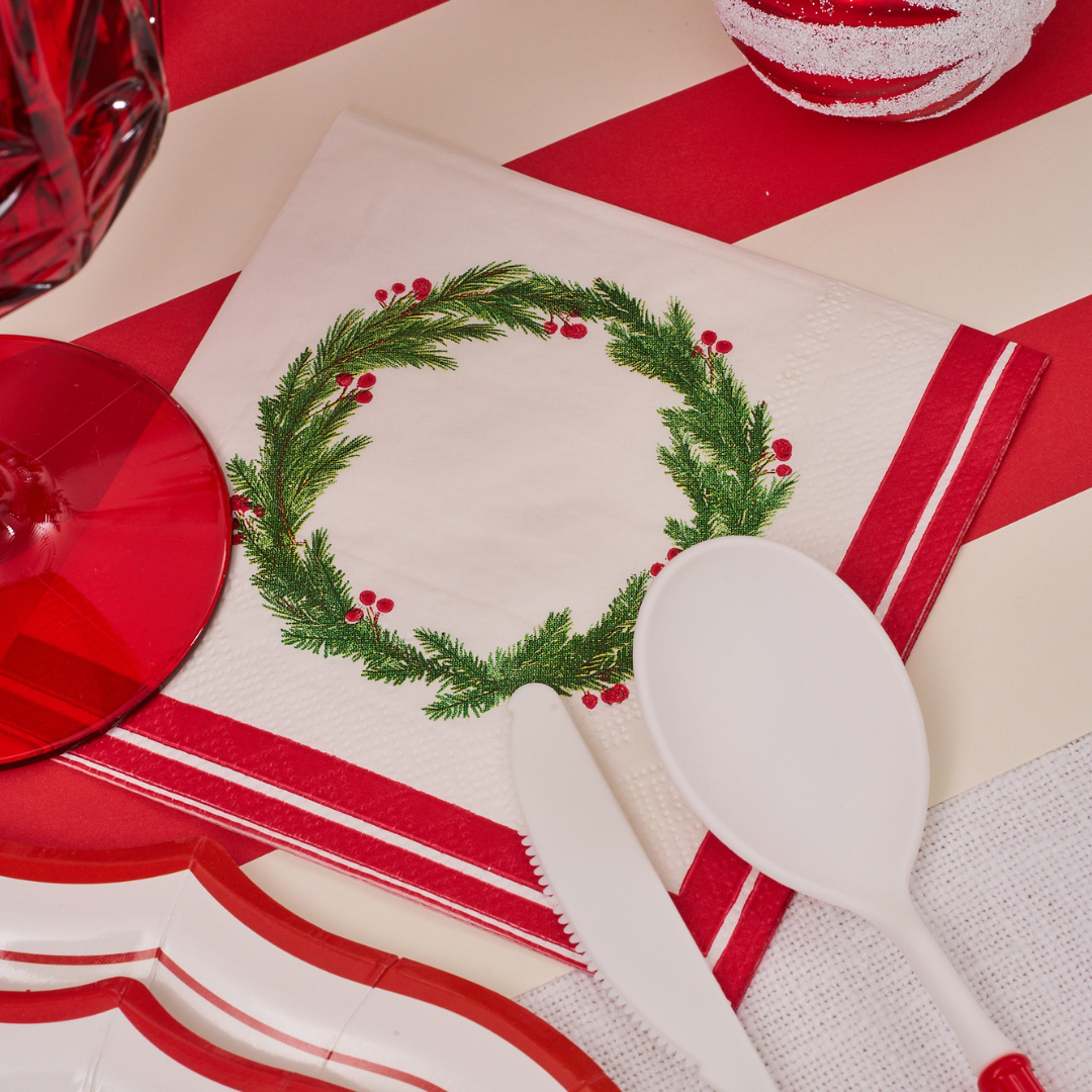 The Merry and Bright Table Setting by Sophistiplate features a green wreath napkin, red glass, white cutlery, and a striped red and white background for a festive Christmas tablescape.