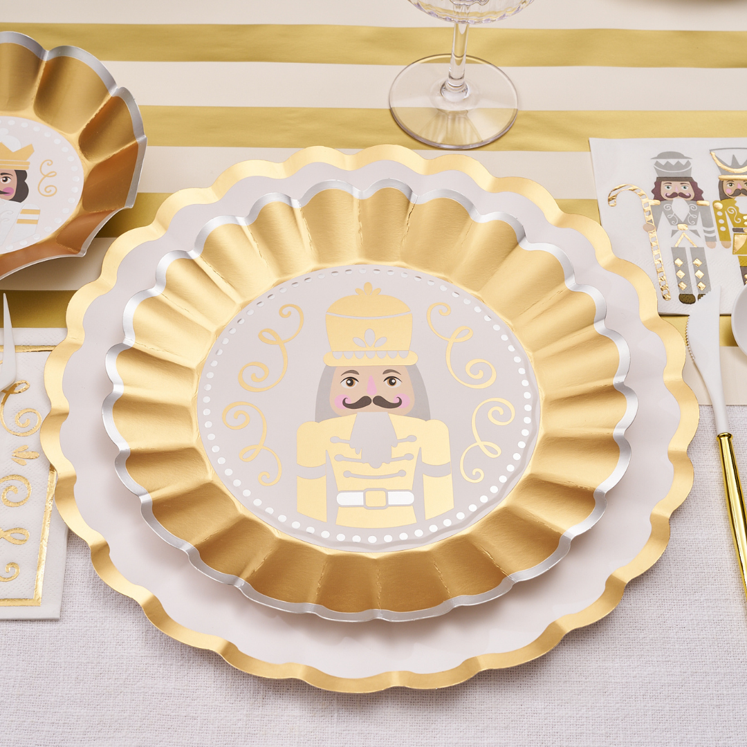 The Gilded Nutcracker Table Setting by Sophistiplate features gold and white scalloped plates with a nutcracker motif, arranged on a striped tablecloth with matching napkins and utensils for an elegant, festive disposable holiday display.
