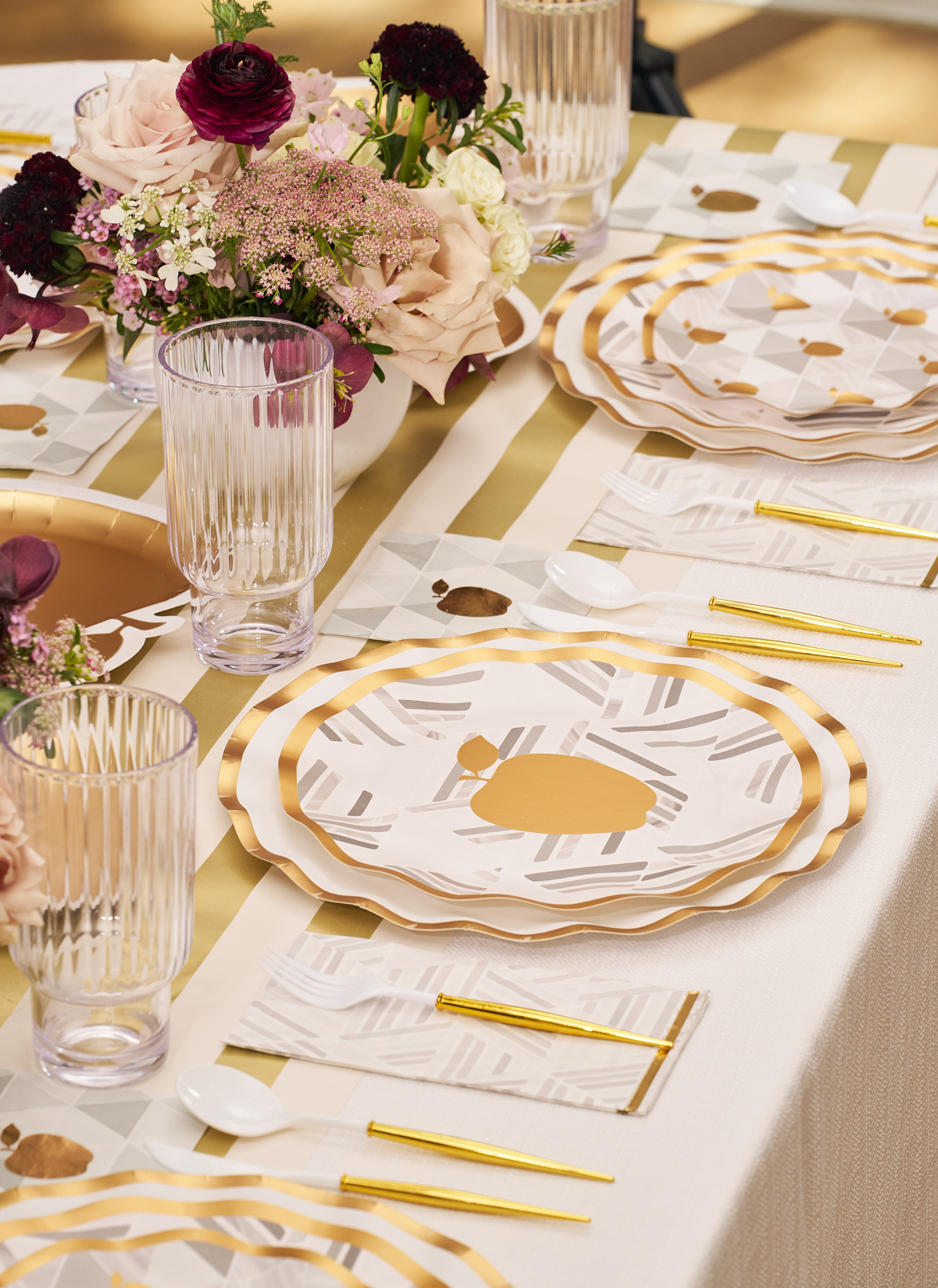 Elevate your Rosh Hashanah celebration with the Golden Apple Table Setting by Sophistiplate. This beautifully coordinated tablescape features a cream and gold striped tablecloth, scalloped disposable plates embellished with an elegant gold apple motif, stylish gold-handled cutlery, ribbed glassware, and floral centerpieces in hues of blush, burgundy, and white—perfect for creating a festive and sophisticated atmosphere.