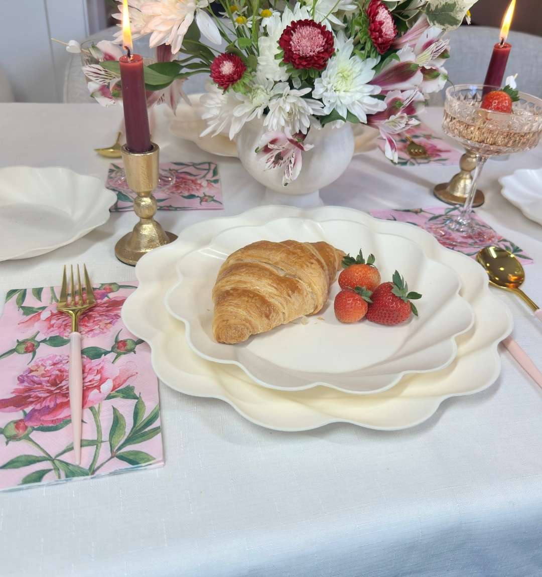 The Eco-Friendly Table Setting - Cream by Sophistiplate features scalloped compostable plates, a croissant, three strawberries, pink floral napkins, pink cutlery, red candles, a gold goblet with a strawberry, and a flower-filled vase.