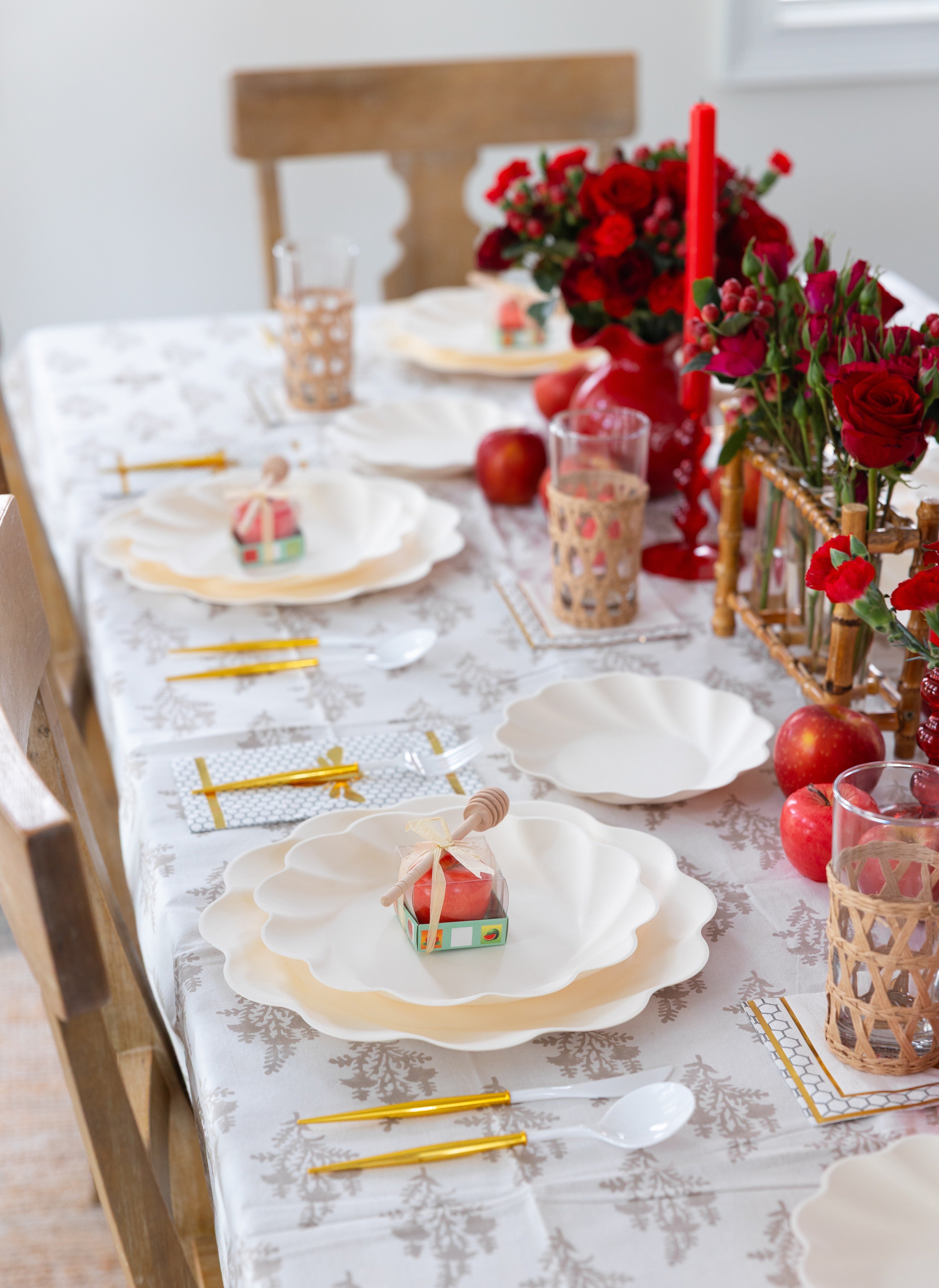 Create a festive table setting for Rosh Hashanah with the “Rosh Tablescape for 16 Guests: Eco Cream Honeybee” by Sophistiplate LLC, featuring elegant white tableware, gold cutlery, apple-inspired accents, honey dippers, red floral arrangements, candles, woven glass holders, and a white patterned tablecloth.