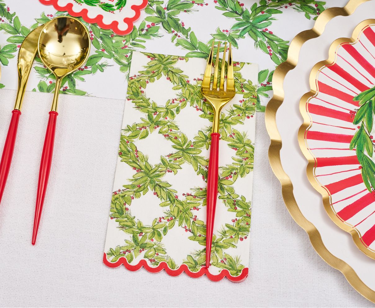 Create a festive look with the Holly Berry Table Setting by Sophistiplate, featuring gold and red utensils, green foliage and berry napkins, and red, white, and gold scalloped disposable plates on a crisp white tablecloth.