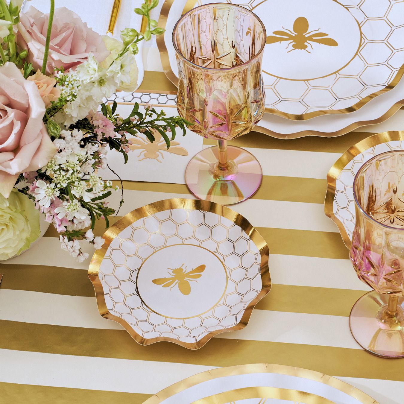 The White Honeybee Table Setting by Sophistiplate features gold-rimmed paper plates adorned with bee and honeycomb designs, complemented by a pink floral arrangement and pink glasses, all beautifully displayed on a gold and white striped tablecloth. This elegant party tableware set adds a sophisticated touch to any celebration while providing the convenience of disposable plates.