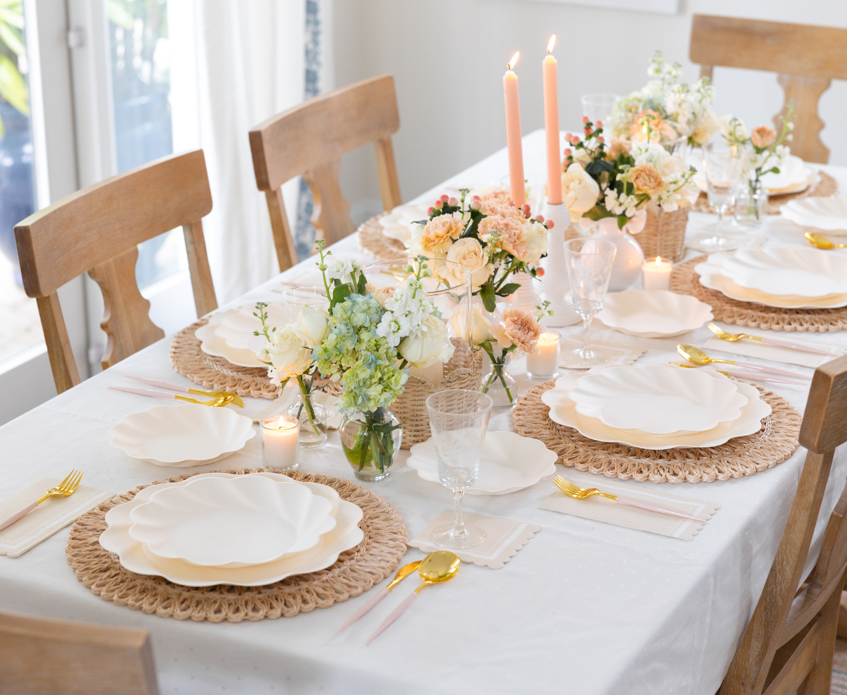 The Eco-Friendly Table Setting-Cream by Sophistiplate features sustainable tableware, gold cutlery, woven placemats, pastel floral centerpieces, candles, and glassware on a white tablecloth with wooden chairs.