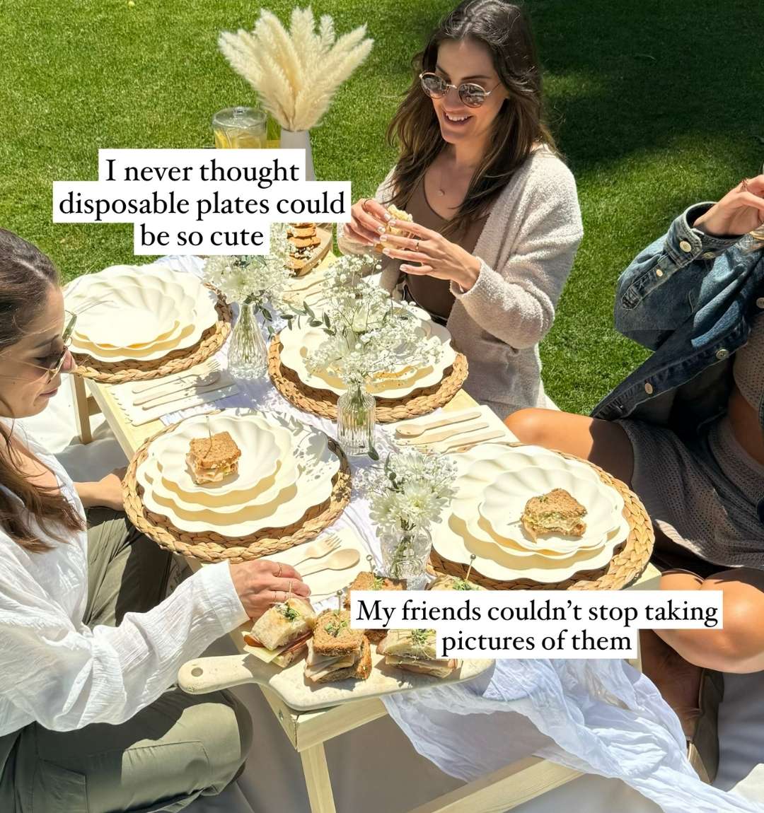 Three women sit outdoors at a picnic table set with Sophistiplate’s Eco-Friendly Table Setting- Cream, plus sandwiches, drinks, and vases of white flowers. Sunlight brightens the scene as two text overlays highlight the cute plates.