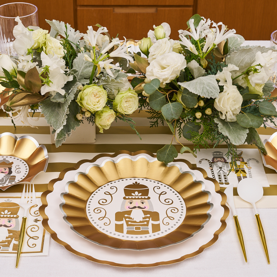 The Gilded Nutcracker Table Setting by Sophistiplate features scalloped-edge plates surrounded by white flowers, greenery, and gold accents for an elegant holiday look with charming disposable tableware.