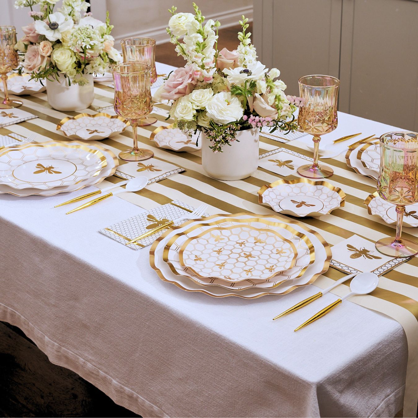 Introducing the White Honeybee Table Setting by Sophistiplate: an elegant arrangement featuring gold rim paper plates and disposable plates, gold utensils, a striped table runner, pink and gold glasses, and floral centerpieces with white and pale pink blooms in white vases—perfect for anyone seeking sophisticated party tableware.