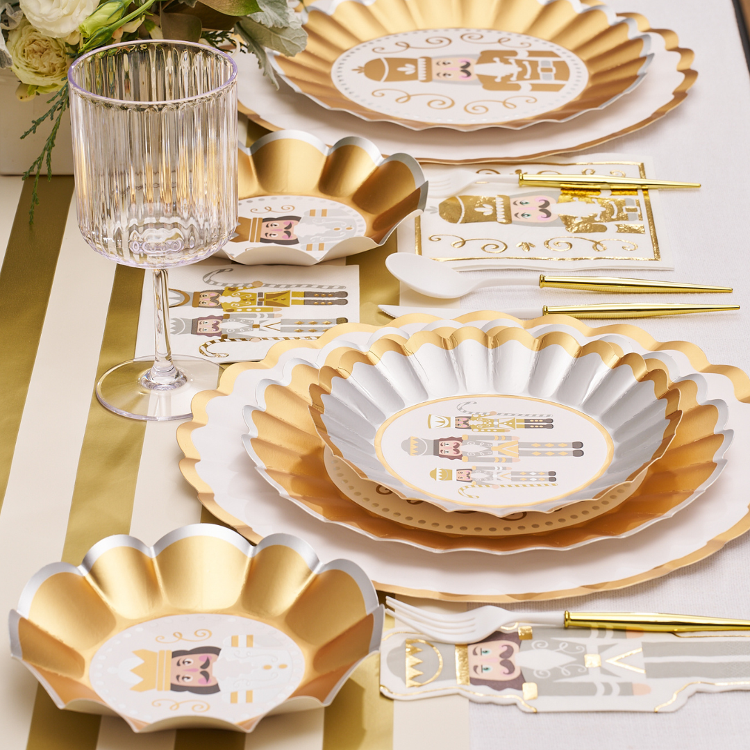 Celebrate the holidays with the Gilded Nutcracker Table Setting by Sophistiplate—scalloped-edge plates, nutcracker napkins, gold utensils, a clear ribbed glass, and floral touches on a striped tablecloth for an elegant disposable setup.