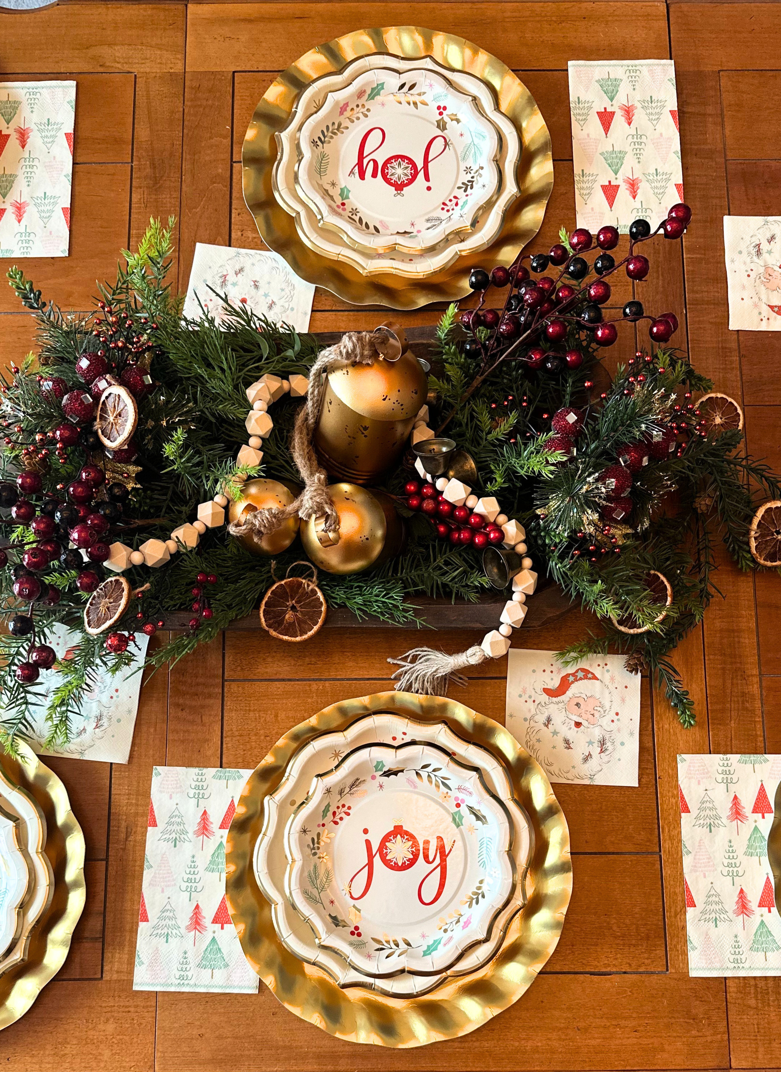 Create a festive table with Sophistiplate LLC’s Vintage Santa: Christmas Table for 16, featuring holiday plates amid evergreens, red berries, candles, ornaments, and themed napkins on a wooden table.
