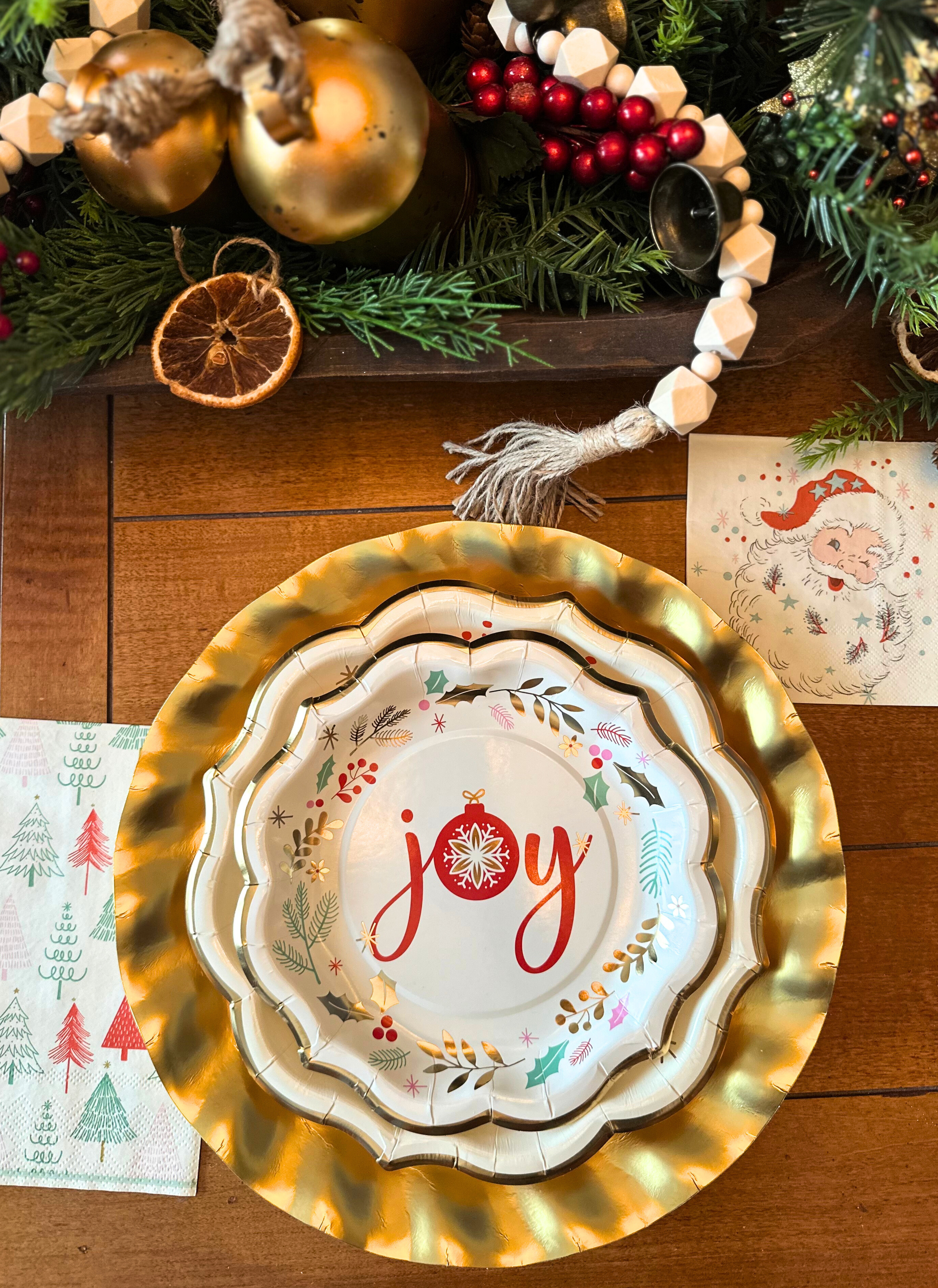 A festive Vintage Santa: Christmas Table for 16 tablescape featuring a designer paper plate with the word joy and a red ornament design, set on a gold scalloped charger beside a Santa napkin, greenery, red berries, and wooden beads.