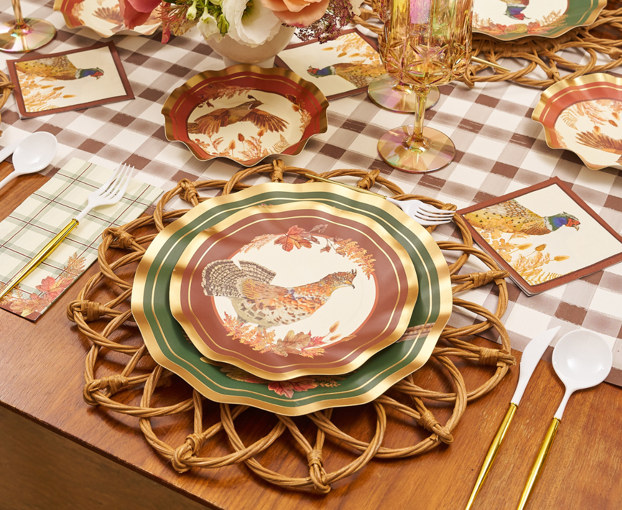 A fall-themed table setting featuring Sophistiplate Woodland Harvest Salad Plates (8pk) with charming bird prints, accompanied by woven placemats, plastic cutlery, napkins, and glasses arranged on a brown plaid tablecloth. A vase of flowers completes the inviting Thanksgiving-inspired atmosphere.