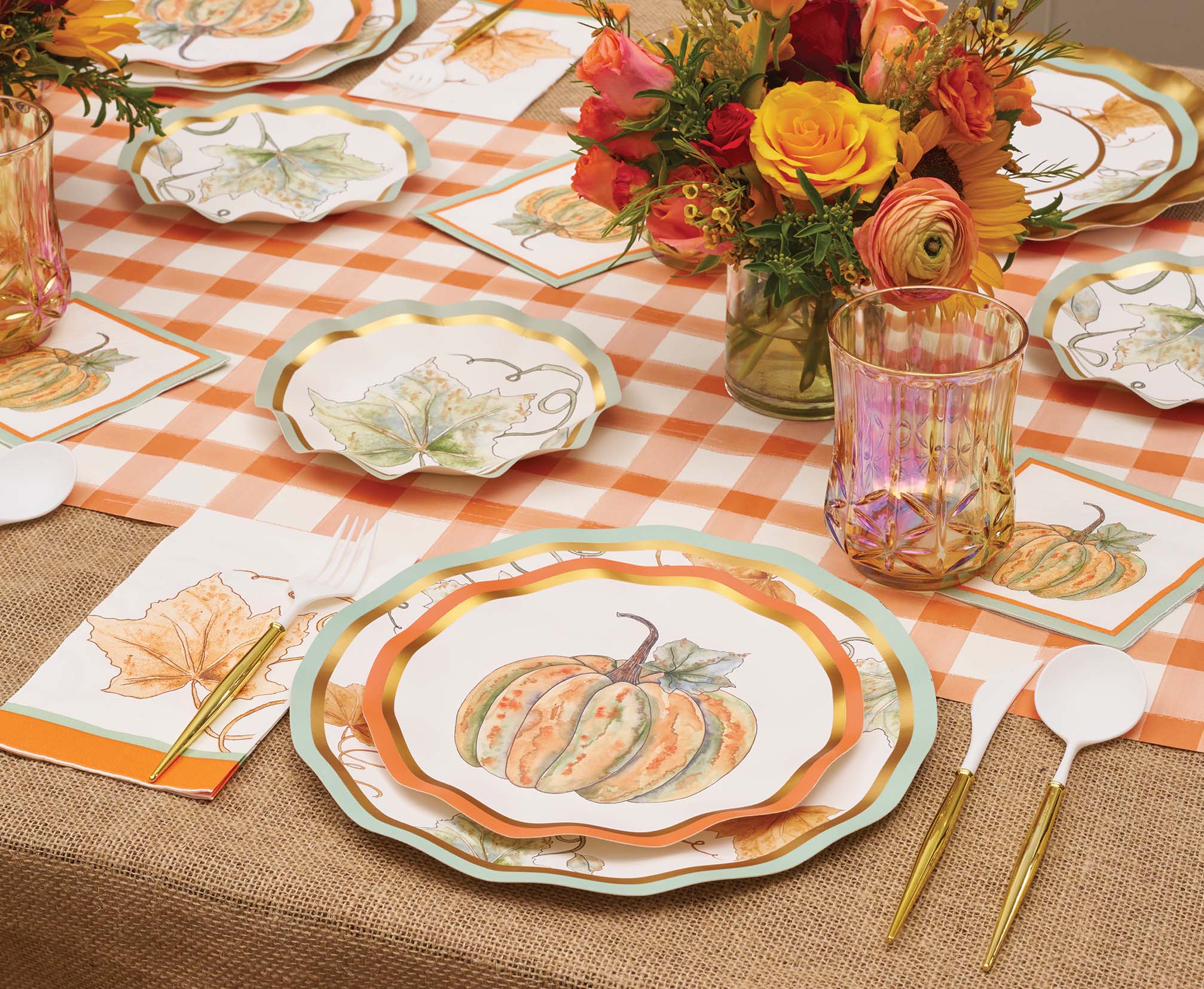 The Bright & Thankful Table Setting by Sophistiplate combines pumpkin harvest plates, gold utensils, glassware, vibrant floral centerpieces, and a checkered tablecloth for a festive Thanksgiving celebration.