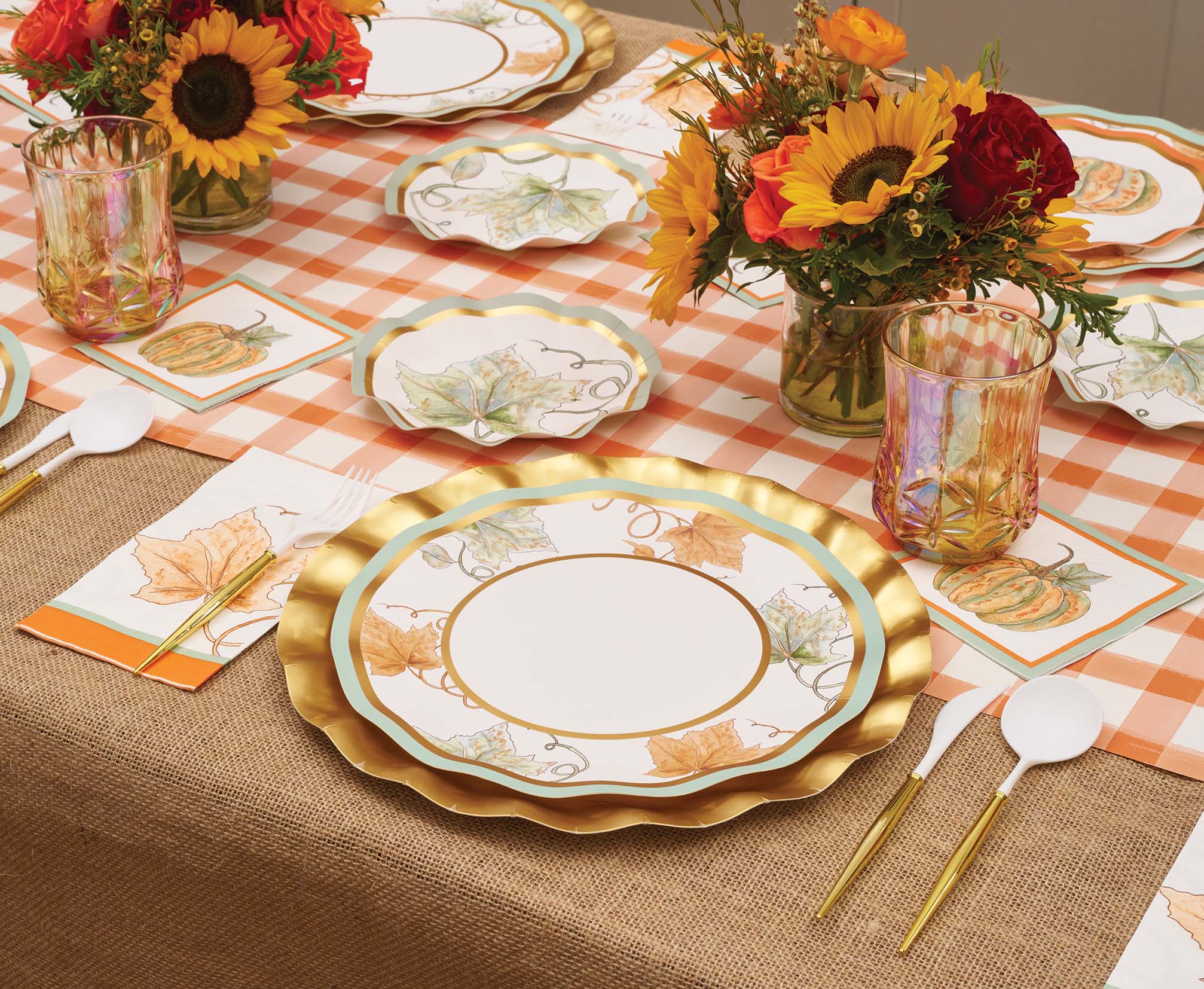 A festive autumn table setting featuring Sophistiplate’s Bright & Thankful Dinner Plates (8pk), each adorned with metallic gold trim, complemented by floral centerpieces, pumpkin-themed disposable plates, elegant gold cutlery, and an orange plaid table runner atop a burlap tablecloth.