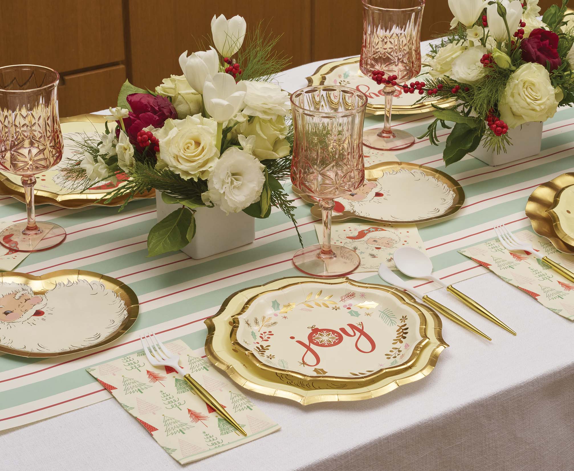 The Vintage Santa Table Setting by Sophistiplate features floral centerpieces, pink crystal glasses, striped runners, JOY plates, Christmas tree napkins, and gold-white cutlery for a festive party table.