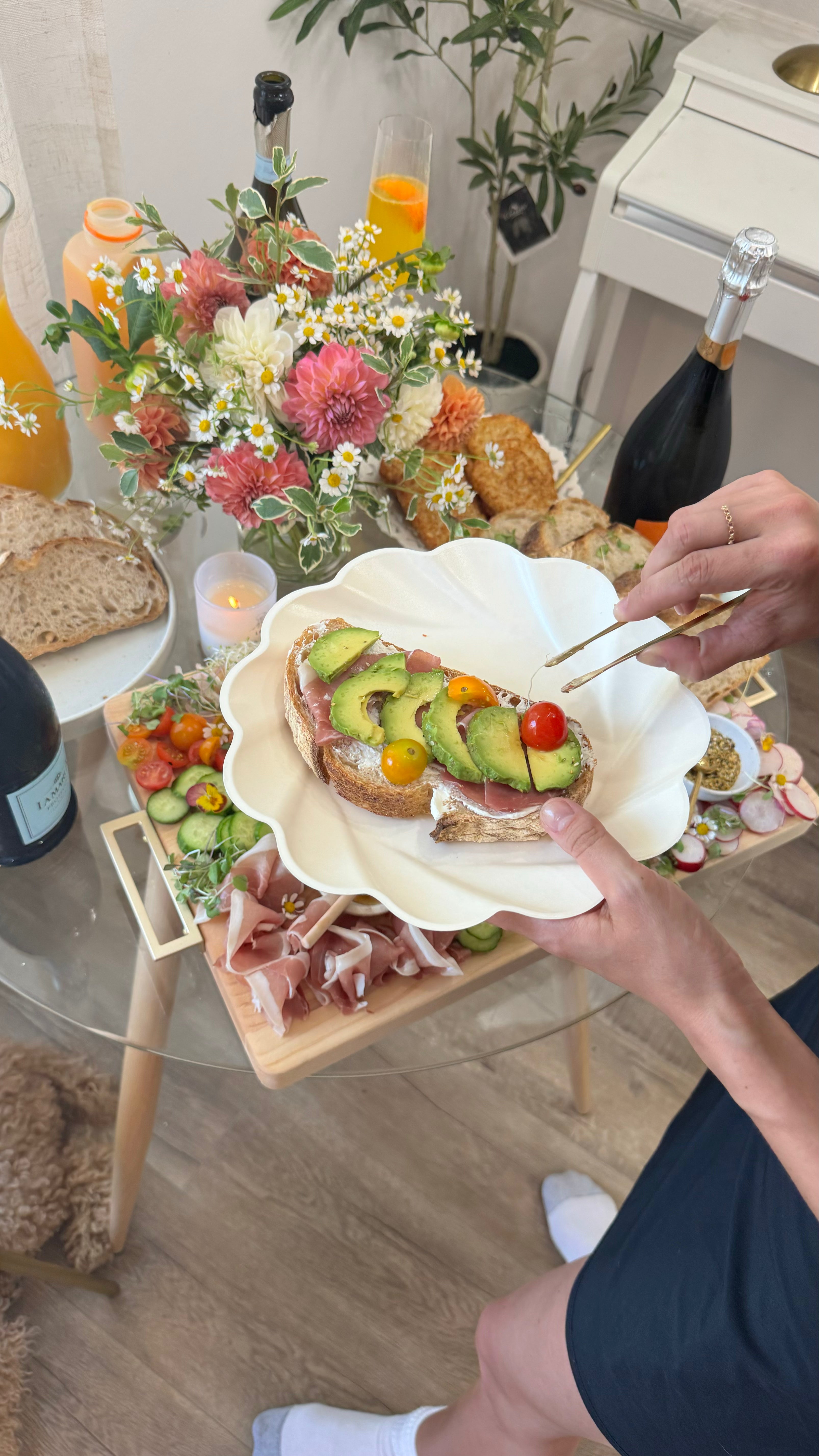 At a vibrant brunch, someone tops avocado toast with cherry tomatoes using Sophistiplate’s Eco-Friendly Table Setting in Cream, surrounded by flowers, wine, juice, fresh bread, charcuterie, and veggies—all served on sustainable tableware.