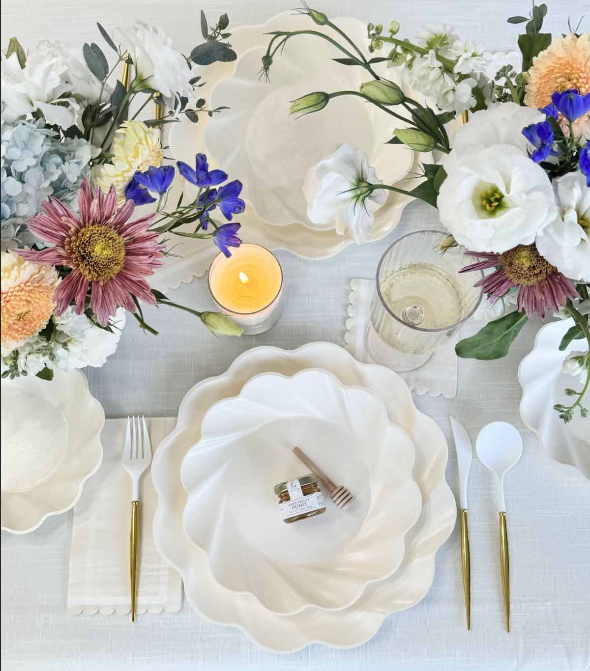 Create an elegant tablescape with Sophistiplate's Eco-Friendly Table Setting in Cream—featuring white scalloped plates, gold and white cutlery, a glass of sparkling water, flowers, candles, and a mini honey jar with a wooden dipper at each setting.