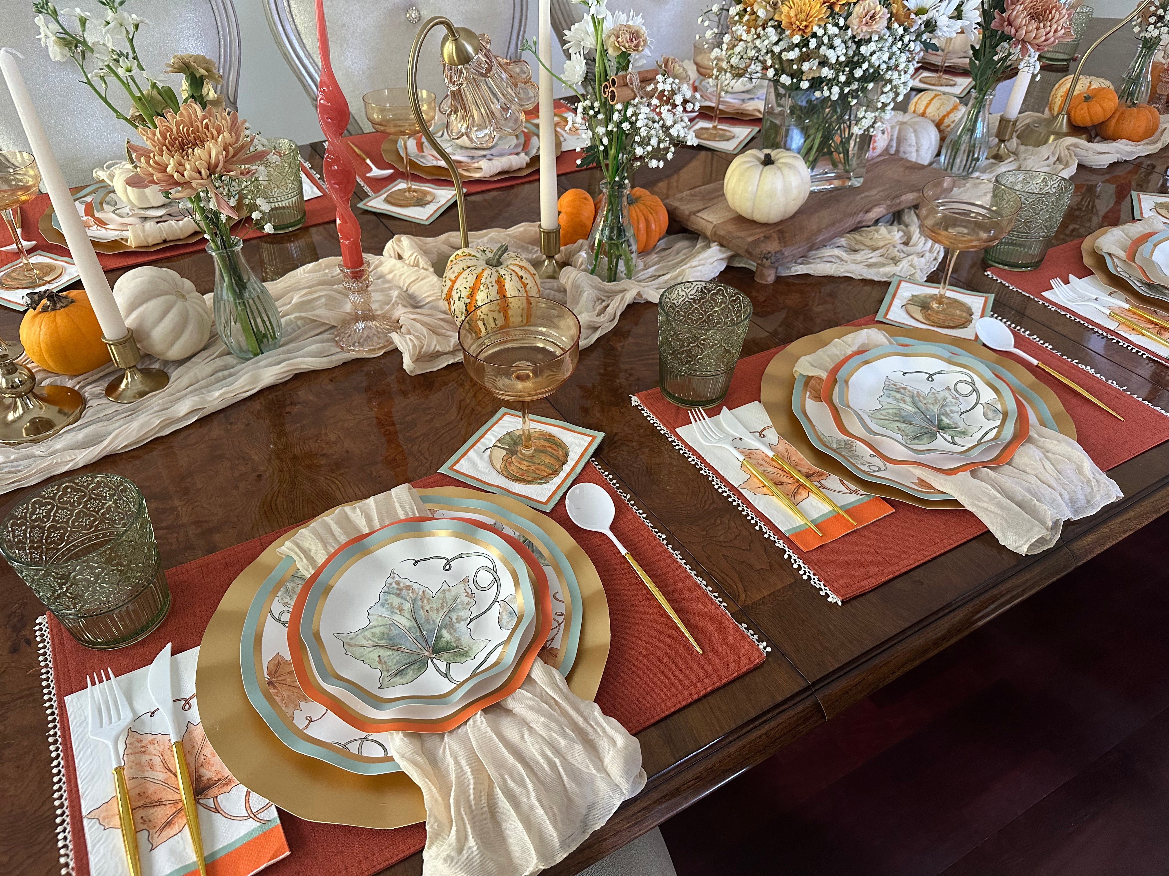 Create a festive autumn tablescape with the Sophistiplate Bright & Thankful Table Setting, featuring pumpkin harvest plates, gold-rimmed glasses, candles, decorative napkins, mini pumpkins, and floral centerpieces in warm orange and beige.
