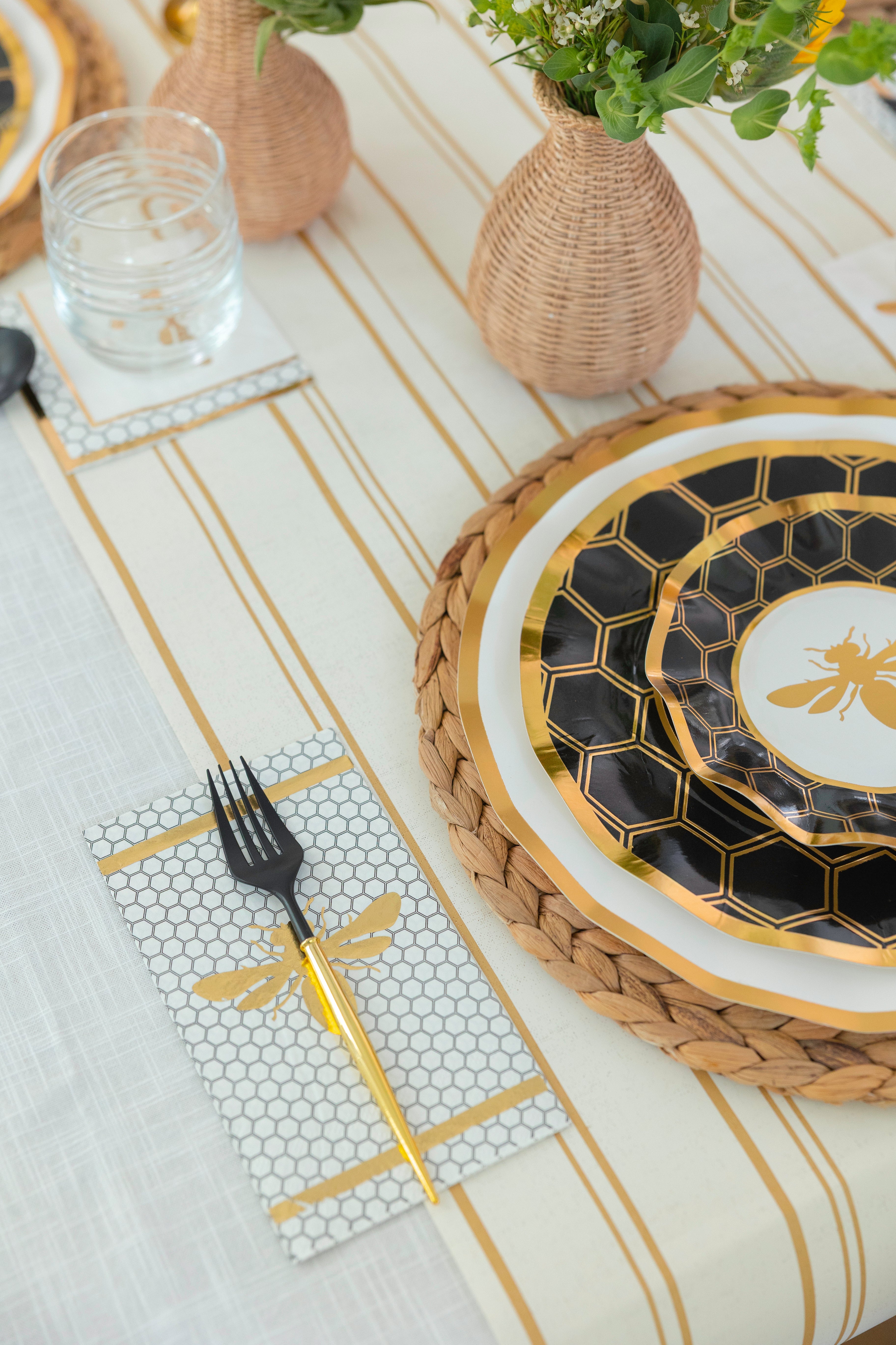 The Honeybee Table Setting by Sophistiplate LLC features gold and black heavy-duty paper plates with a bee design, a gold fork on a honeycomb-patterned napkin, woven vases with flowers, and a striped tablecloth.