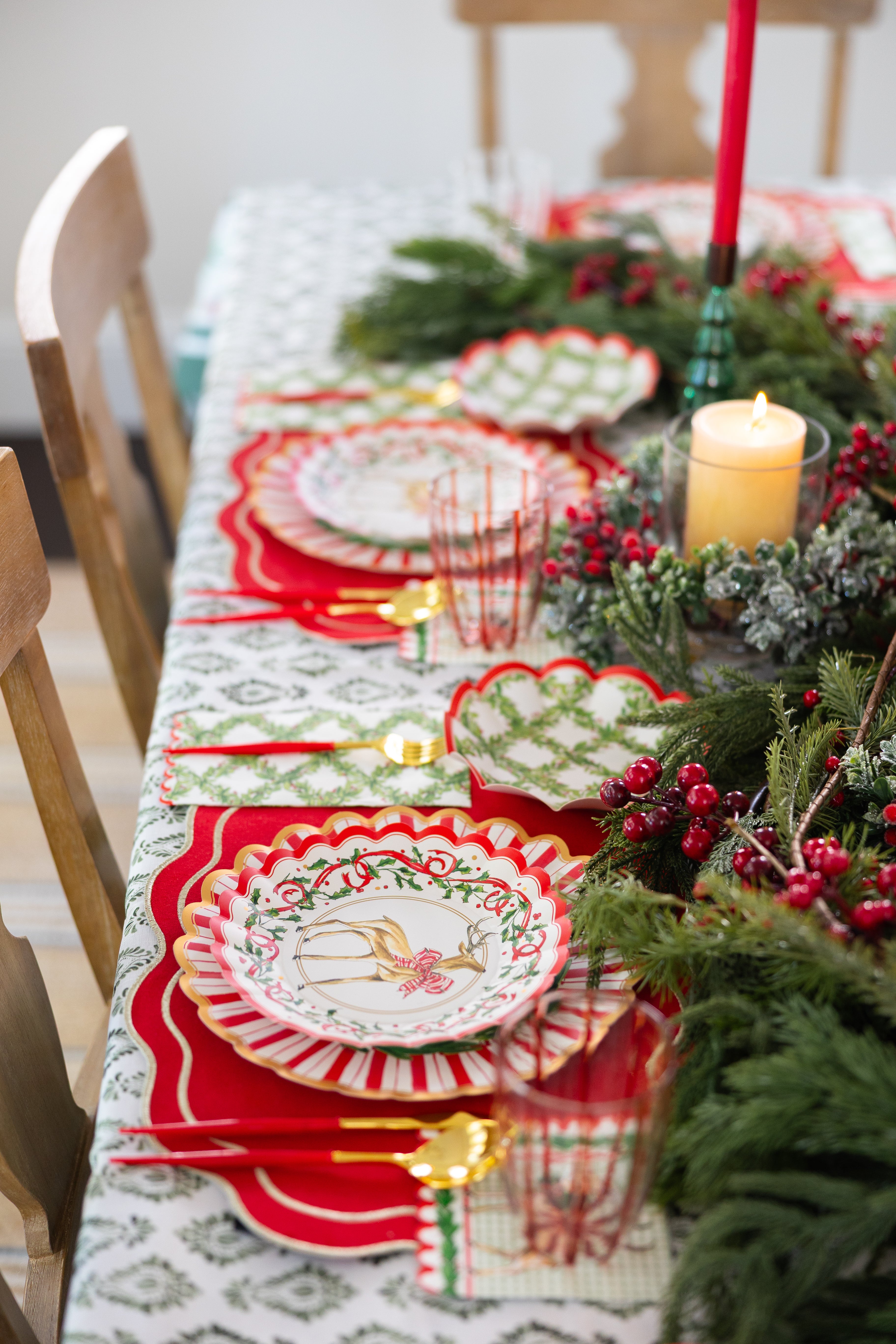 Create a festive holiday tablescape with the Sophistiplate Holly Berry Table Setting—designer disposable plates, gold cutlery, red-and-green glassware, a glowing candle, and a greenery centerpiece atop a decorative tablecloth.