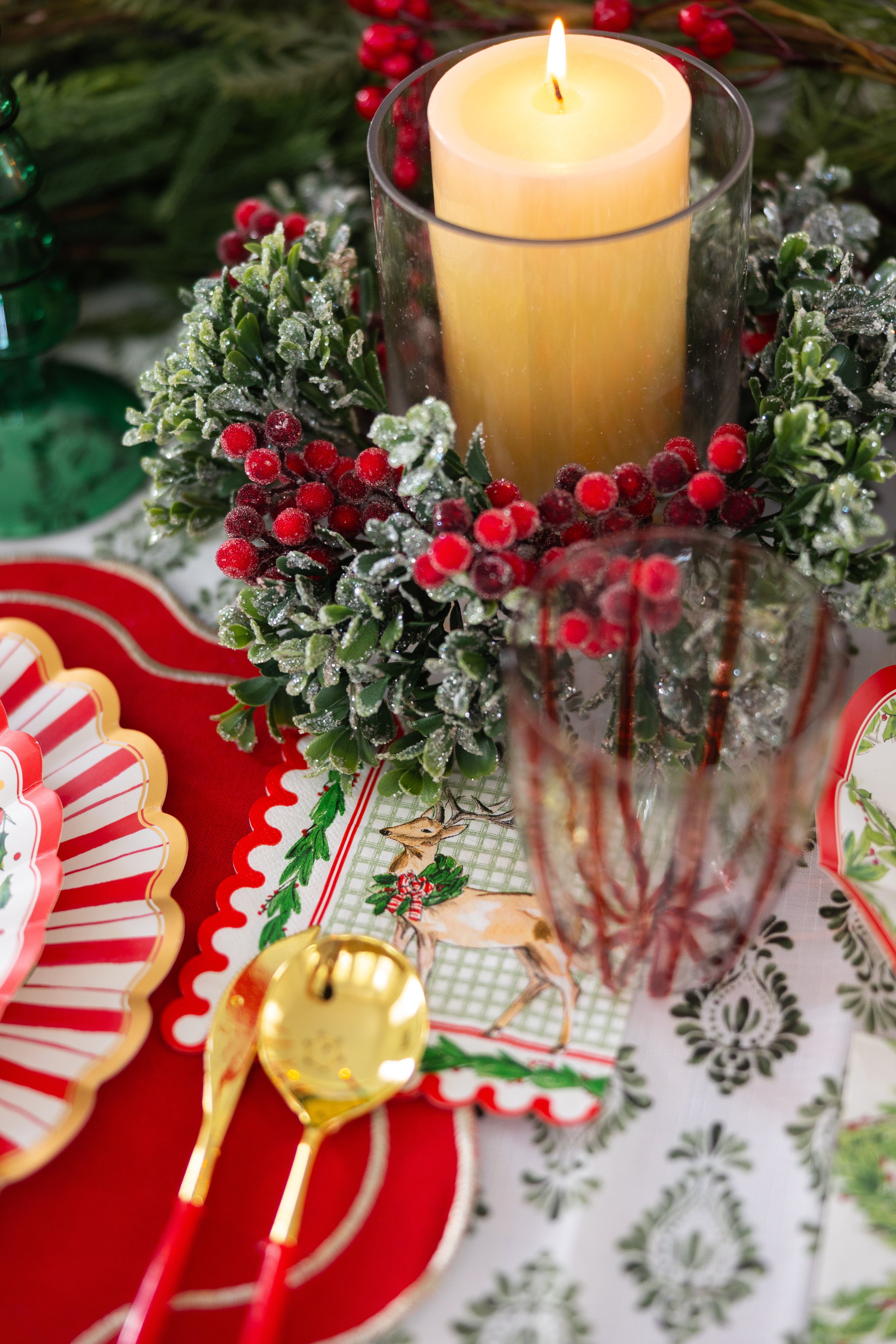 The Holly Berry Table Setting by Sophistiplate features a festive tablescape with a pillar candle in a glass holder, frosted greenery, red berries, gold and red cutlery, designer disposable plates, and reindeer placemats.