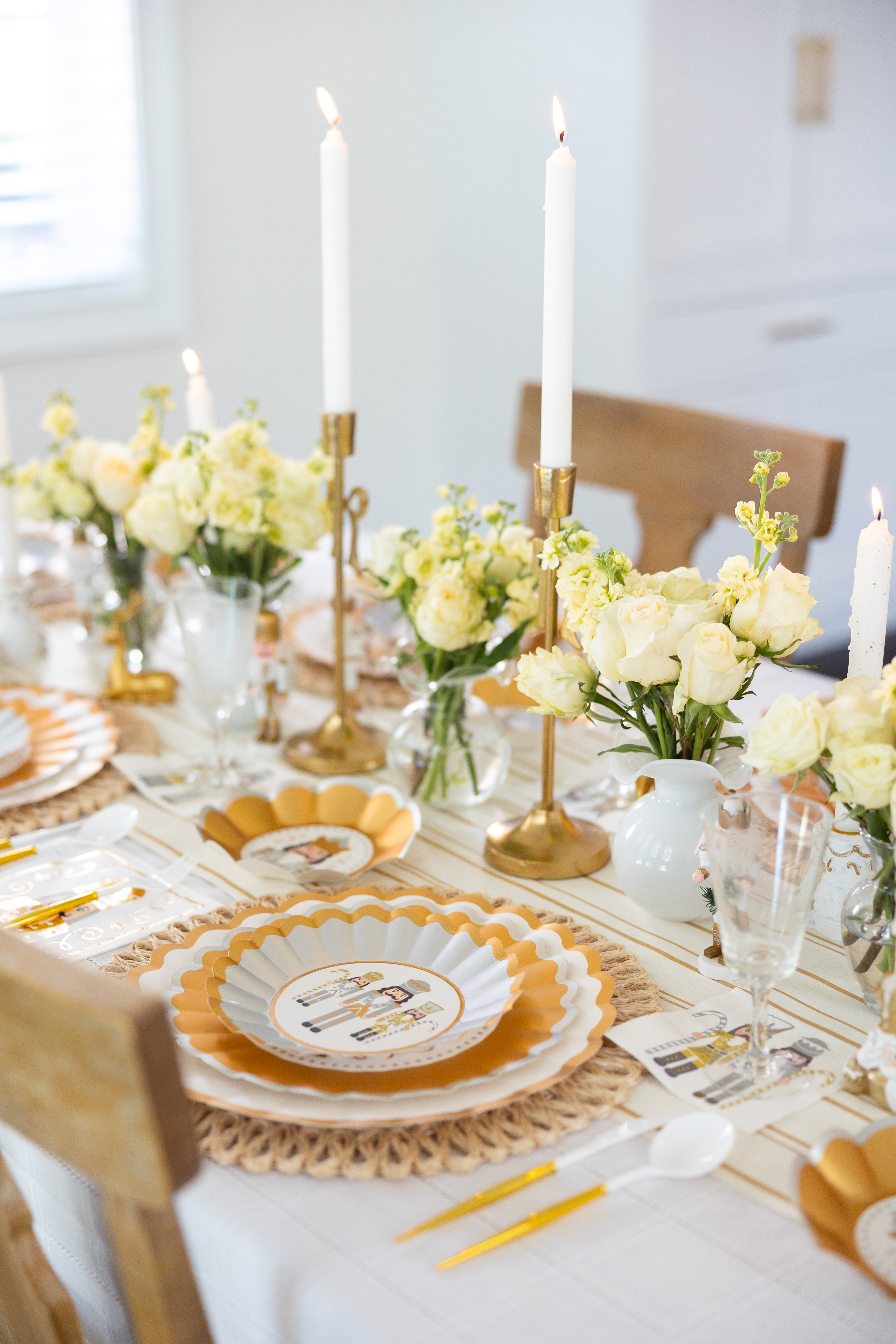 A bright, elegant table set with Sophistiplate’s Gilded Nutcracker Table Setting features scalloped plates, gold flatware, white candles in gold holders, and vases of white and yellow flowers on a crisp white tablecloth.