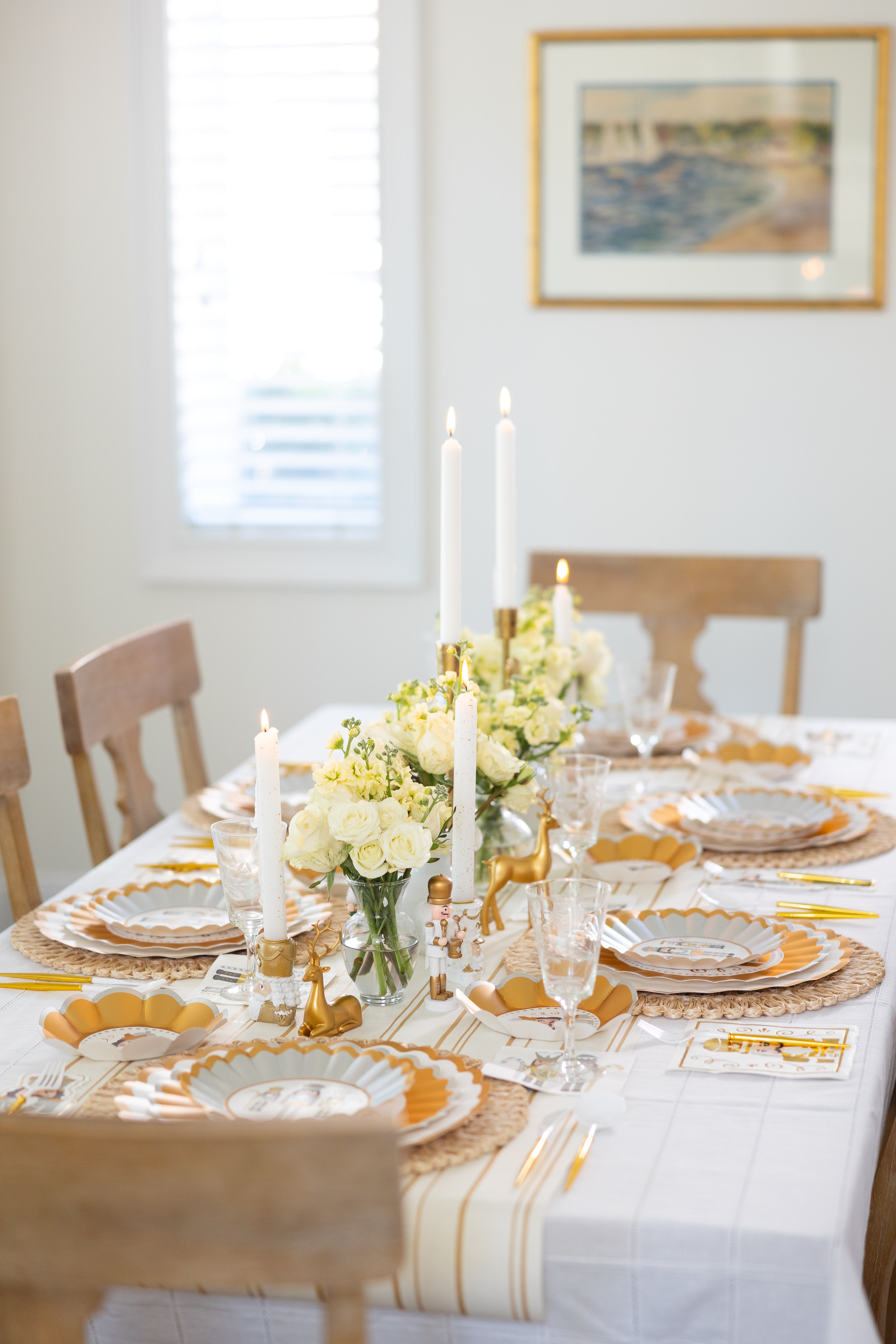 The Gilded Nutcracker Table Setting by Sophistiplate features scalloped edge party plates, glassware, white candles in gold holders, and white and yellow floral centerpieces on a dining table with four wooden chairs in a bright room.