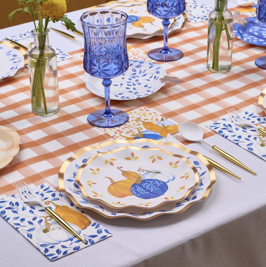 The Harvest Blues Table Setting by Sophistiplate features pumpkin-themed disposable plates, gold utensils, blue patterned napkins, blue glass goblets, and yellow flower vases on a peach and white checkered tablecloth.