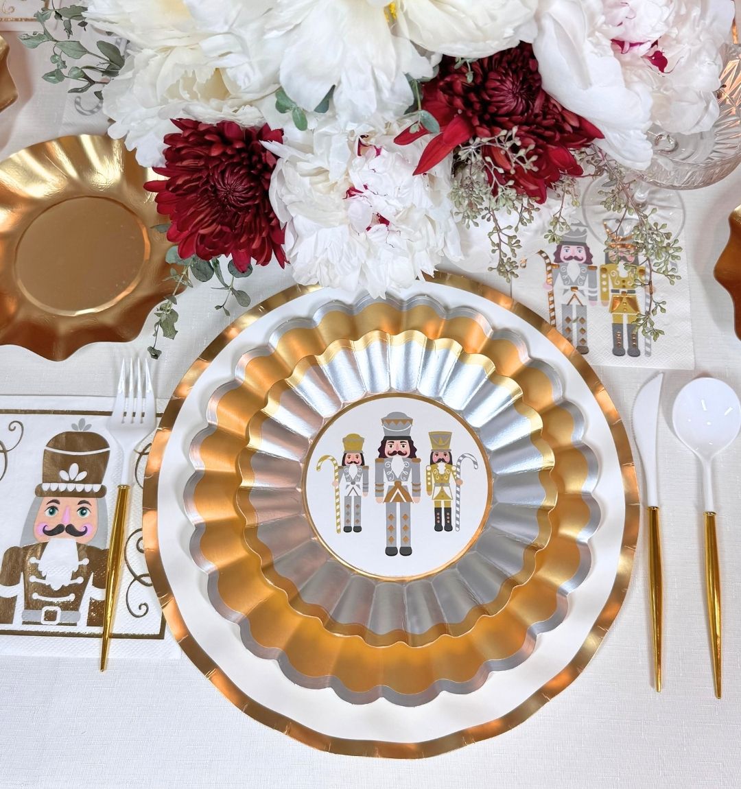 A Gilded Nutcracker: Christmas Table for 16 setting featuring scalloped gold and silver disposable party plates with nutcracker illustrations, matching napkins, white cutlery with gold handles, and a floral centerpiece.