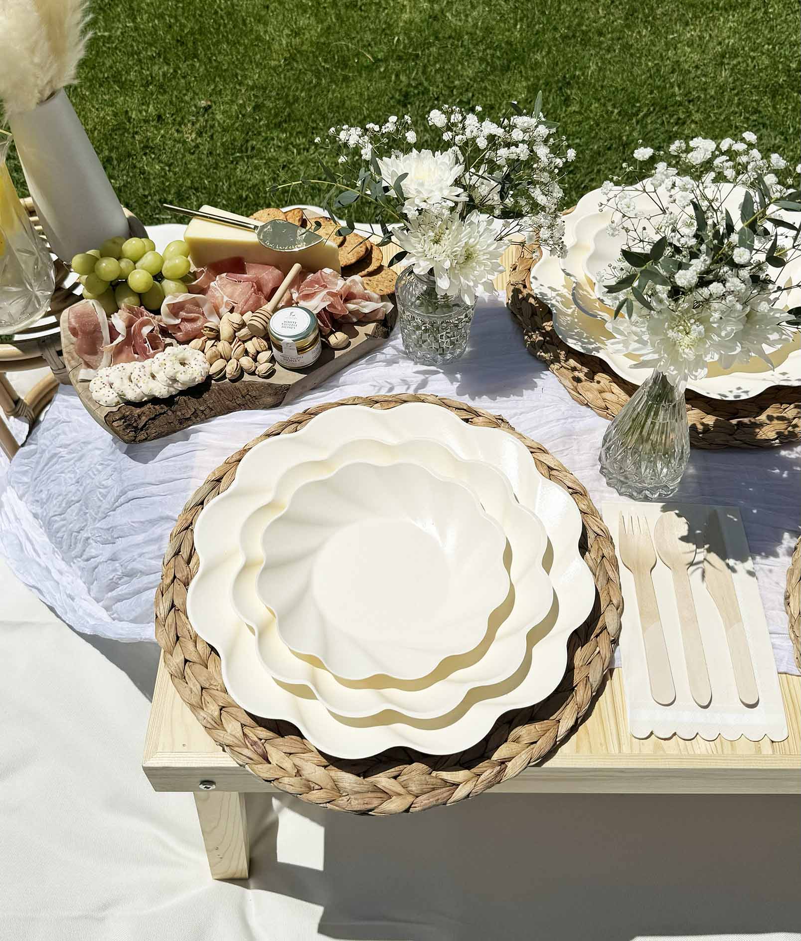 The Eco-Friendly Table Setting- Cream by Sophistiplate stylishly decorates a grassy picnic with compostable party plates, woven placemats, wooden cutlery, glass vases of white flowers, and a charcuterie board of grapes, meat, cheese, and nuts.