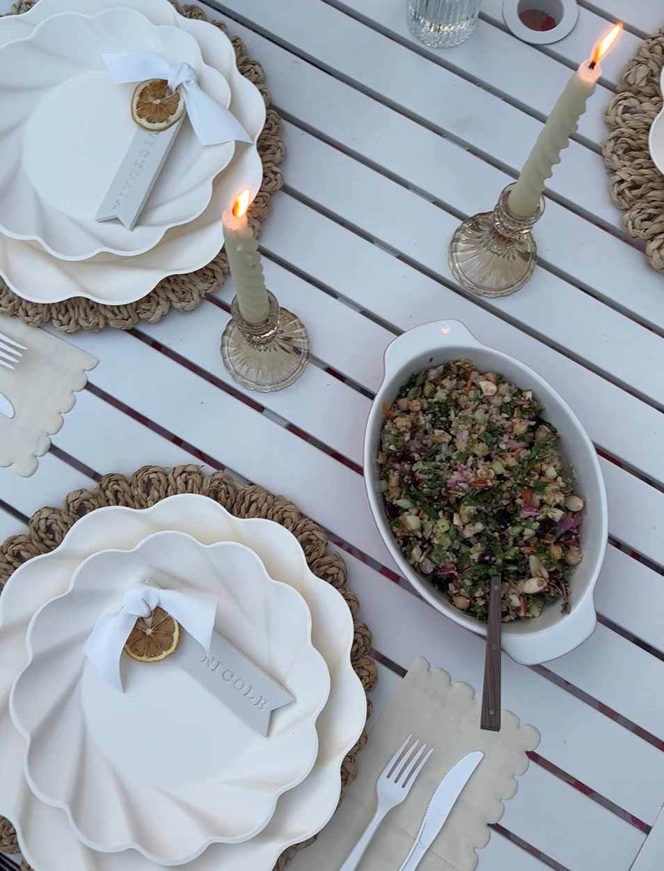 A white outdoor table set with Sophistiplate Simply Eco Dinner Plates in Cream, woven placemats, name tags tied with ribbon, two lit candles in glass holders, and a serving dish of chopped salad with a serving spoon.