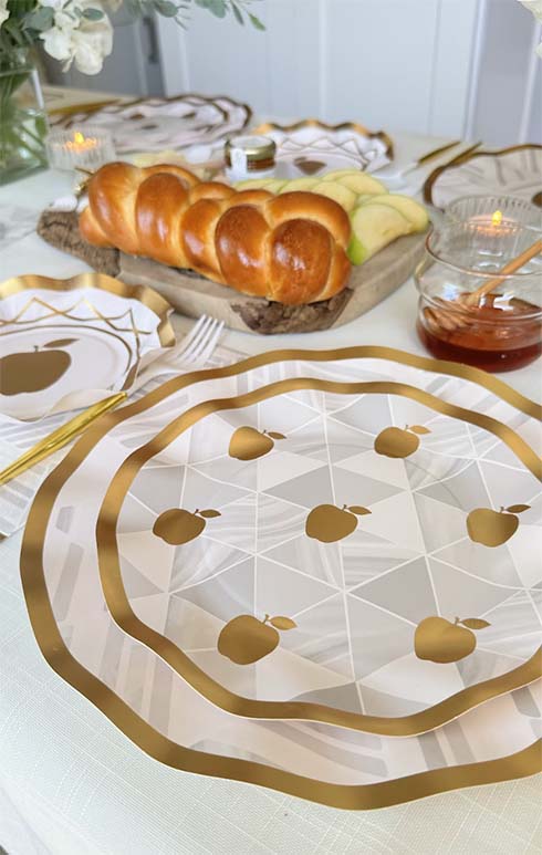 The Sophistiplate LLC “Rosh Tablescape for 16 Guests: Golden Apple” set, complete with designer disposable plates featuring gold-accented apple-themed designs, honey in a jar, sliced apples, challah bread, and decorative napkins on a white tablecloth, creates the perfect Rosh Hashanah table setting.