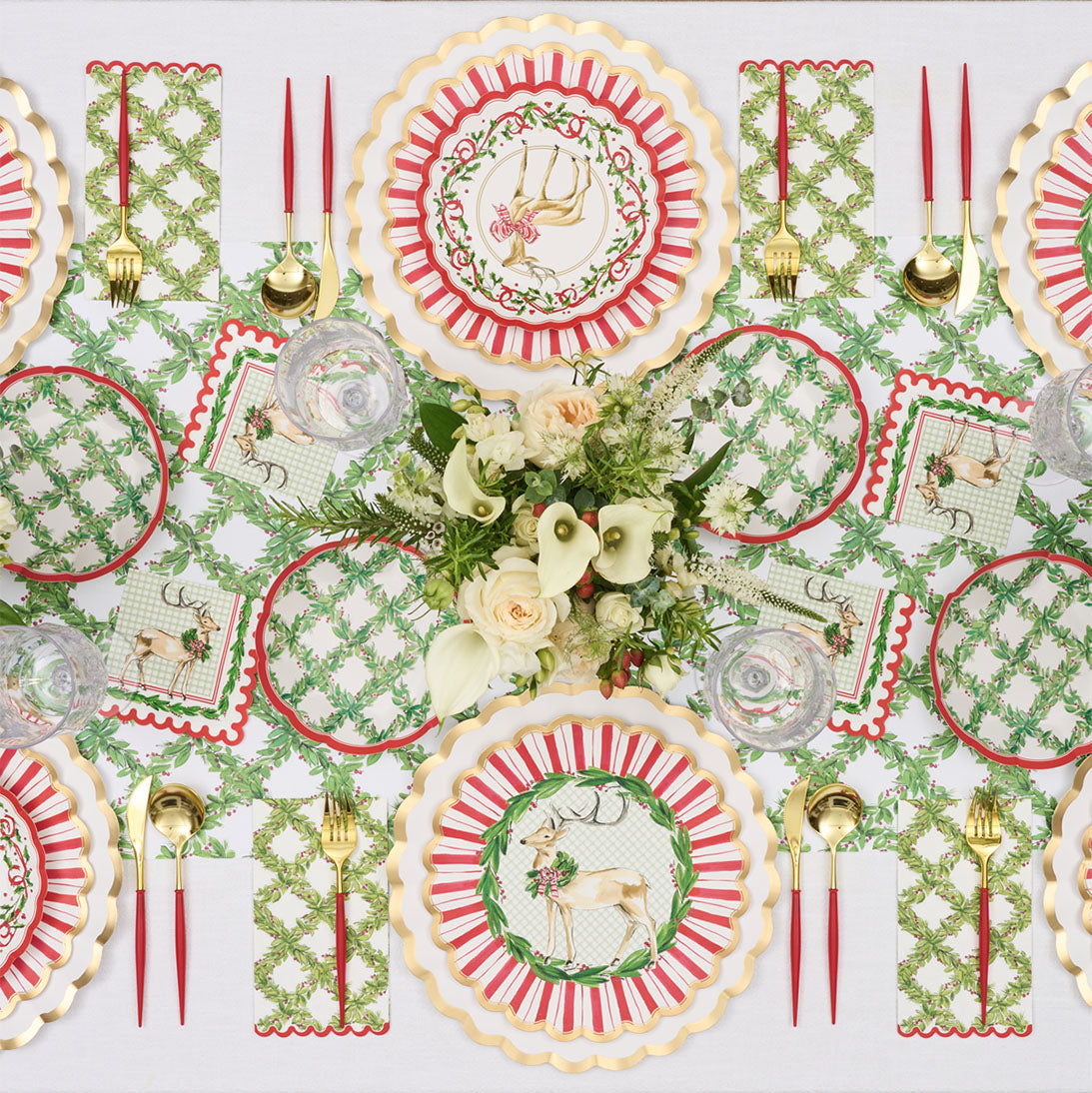 A festive Christmas table features red, gold, and green reindeer and monogram plates, gold utensils, crystal glasses, and a centerpiece of white calla lilies and roses on the Rosanne Beck Holly Berry Trellis Table Runner (20x12